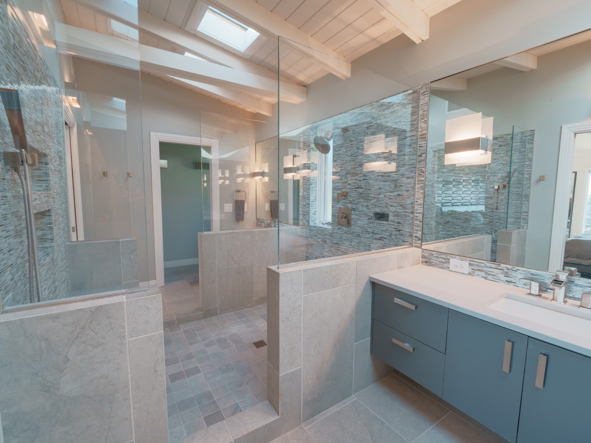 Skylights above the vaulted ceiling bring in additional daylight, while a floating vanity in a soft blue tone with sleek hardware sits beneath a wide mirror and modern sconces.