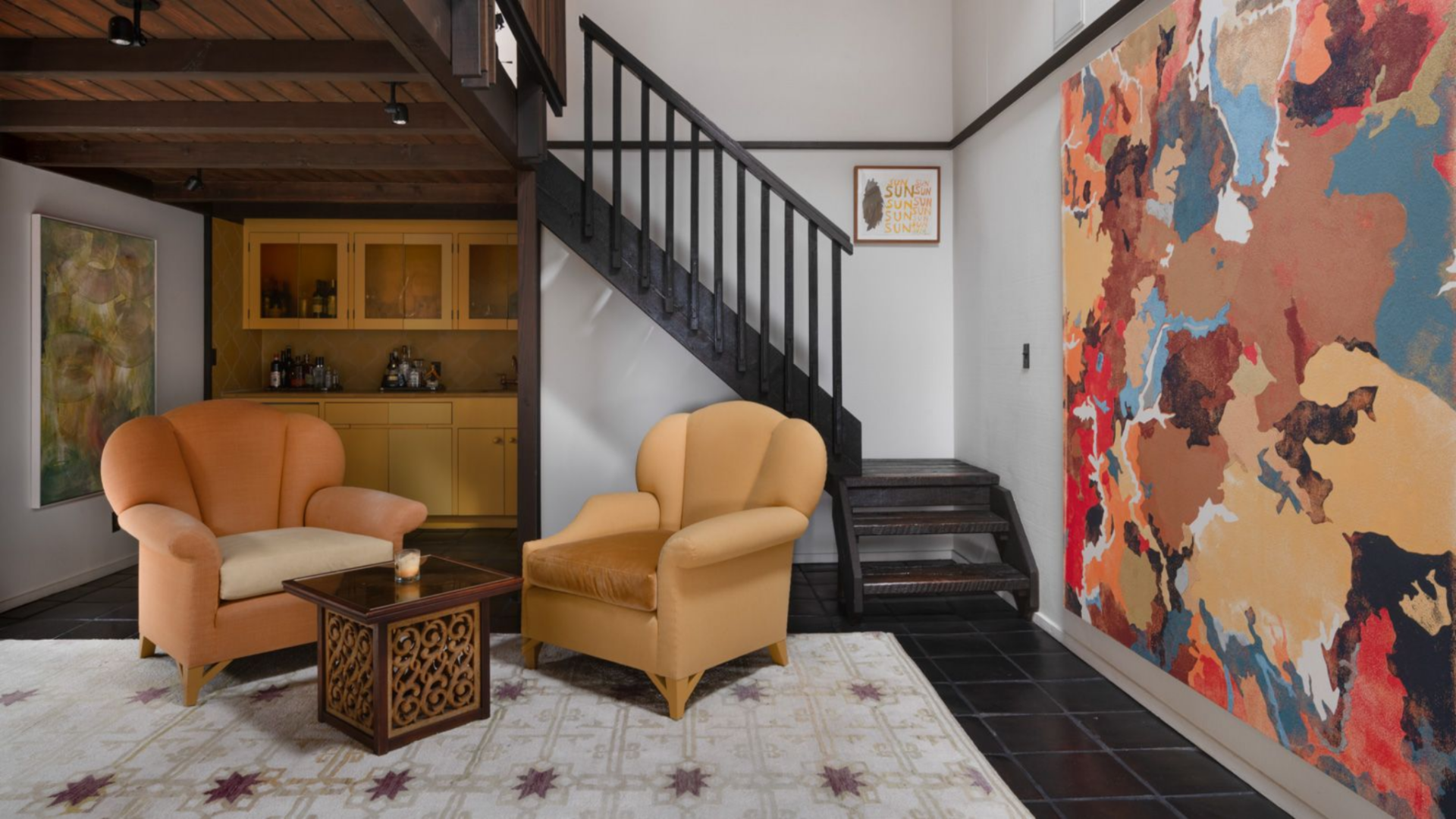 Just off the lounge, a wet bar is tucked beneath the staircase with built-in cabinetry in a muted yellow and glass-front uppers displaying bottles and barware.
