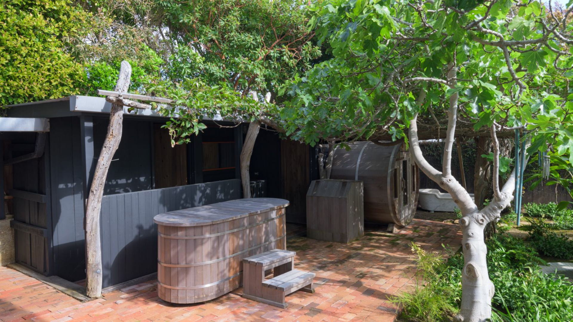 Just a few steps away, a barrel sauna and cold plunge are set on a brick-paved section of the yard, surrounded by trees, creating a compact wellness area within the landscape.