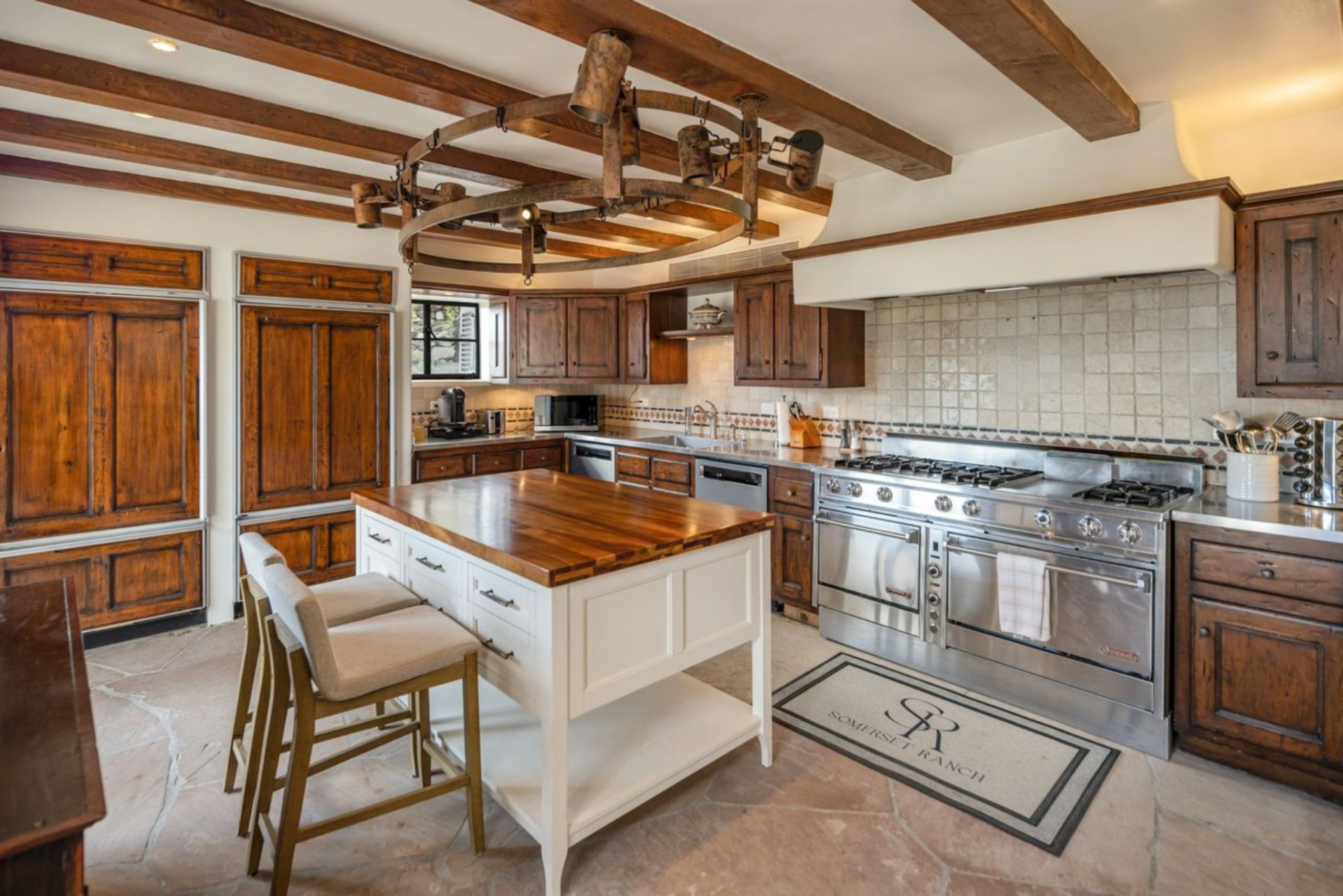 In the main kitchen, exposed ceiling beams run overhead, centered by a circular iron light fixture, while a central island with a butcher block top adds a practical workspace between the two zones. 