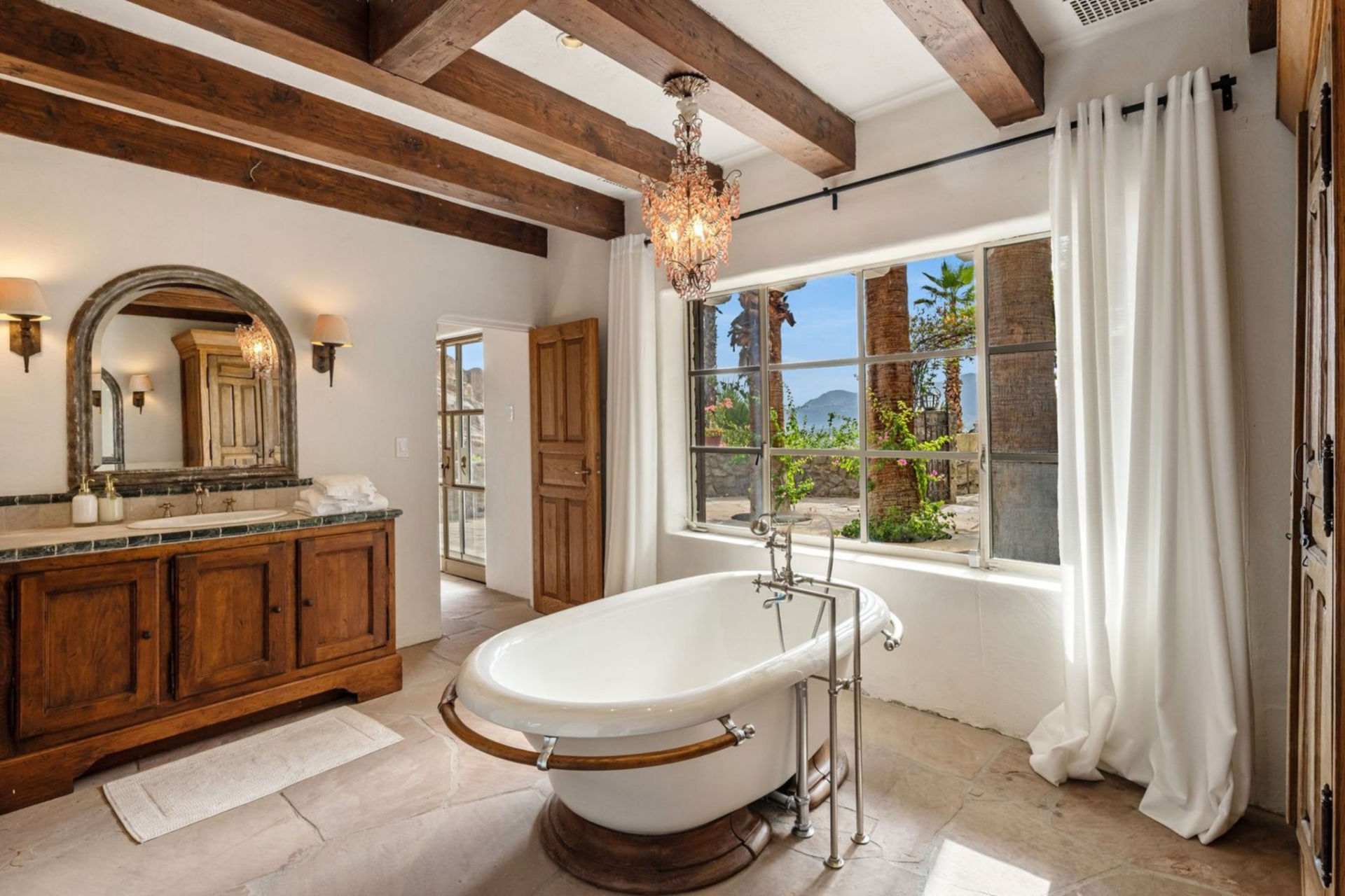 Through a doorway, the bathroom is lined in wood cabinetry and stone flooring, with a freestanding soaking tub placed in the center.
