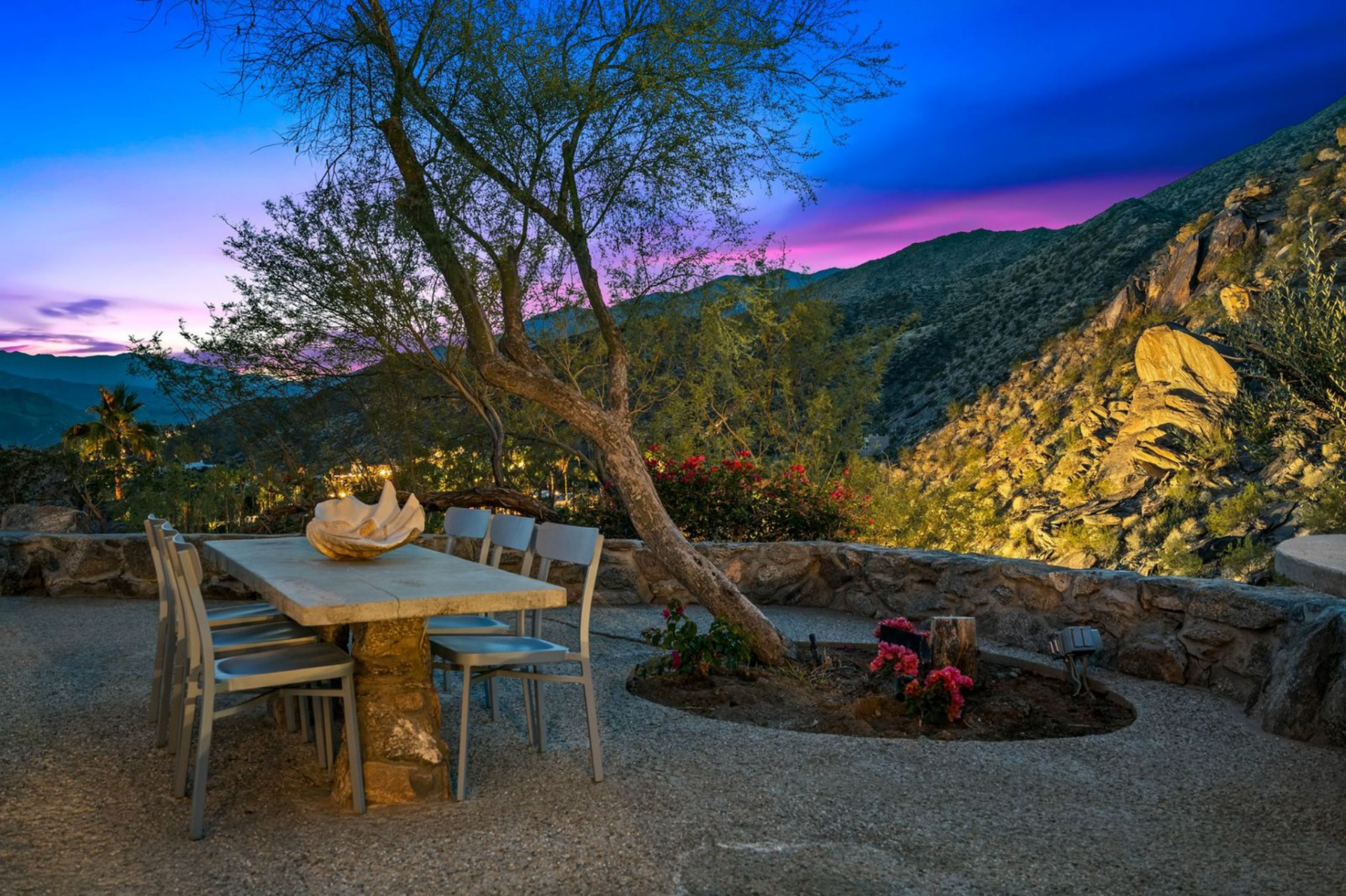 In the outdoor dining area, a stone table is set beneath a tall tree, surrounded by a low rock wall that frames the mountains.
