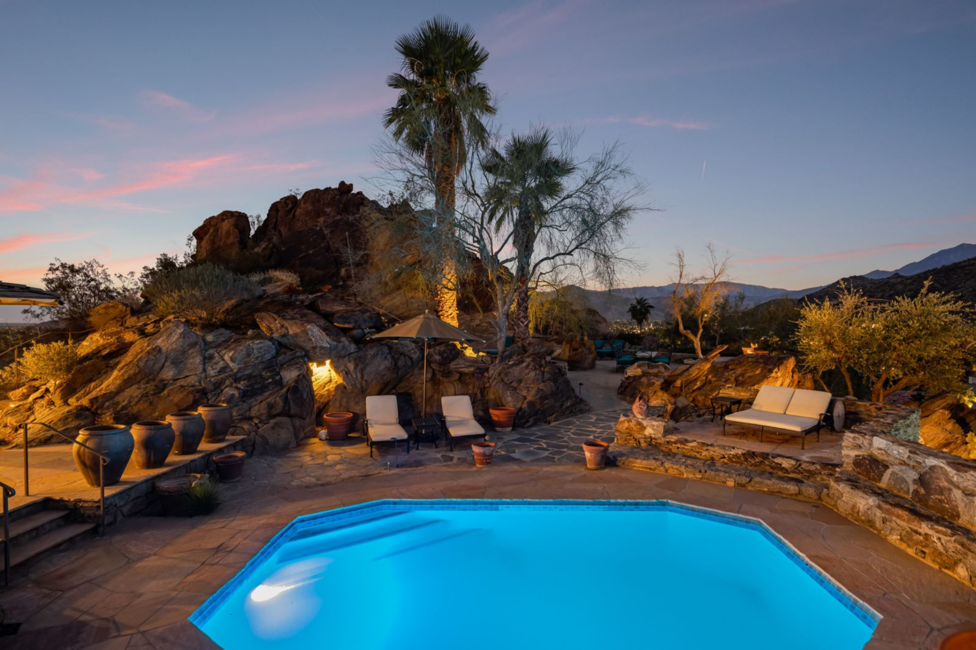 The pool sits within a series of terraced stone decks, surrounded by boulders, palm trees, and lounge areas.