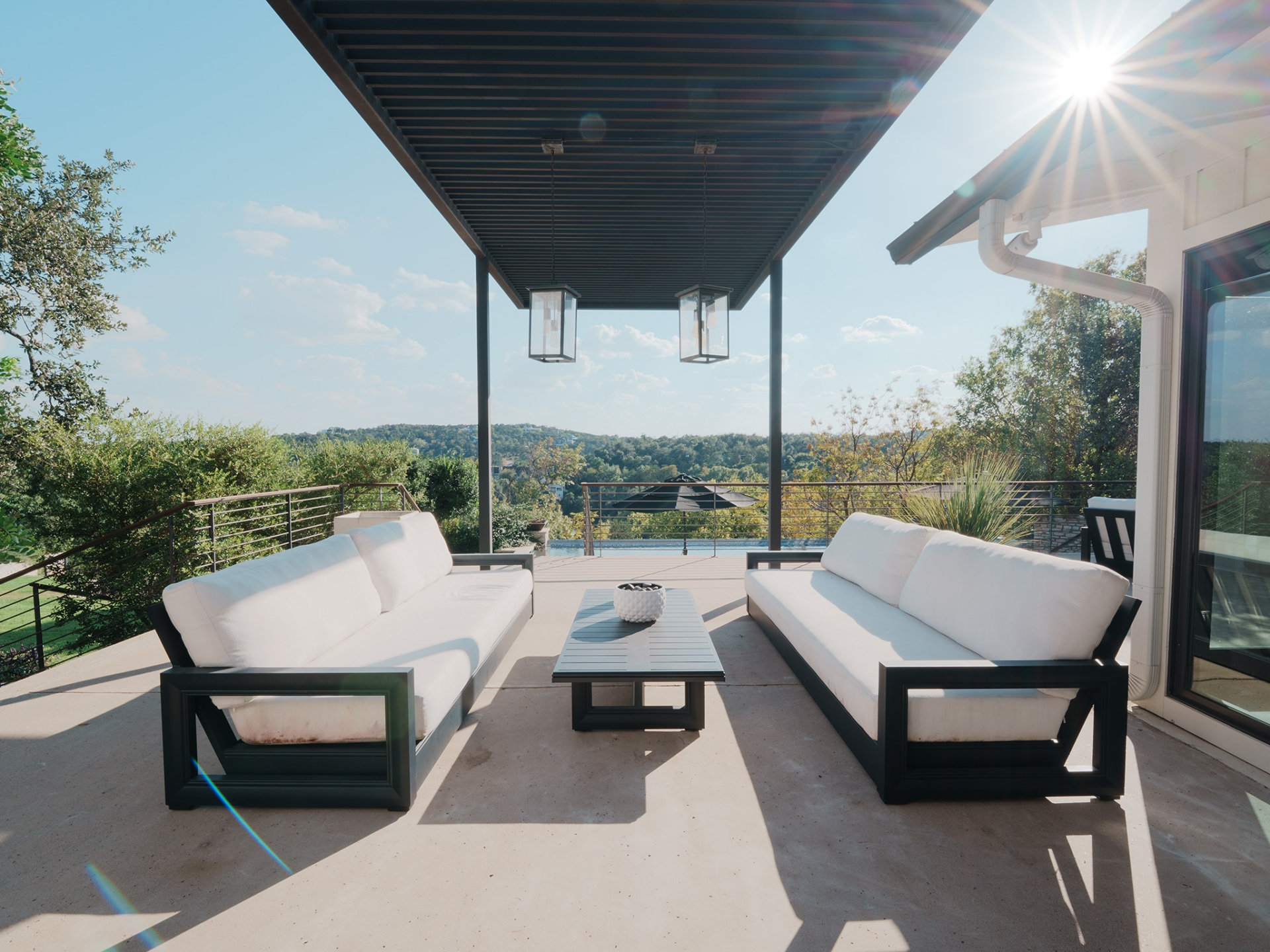 The covered lounge sits just off the main living areas, framed by a sleek metal pergola with two oversized lantern pendants suspended overhead. 