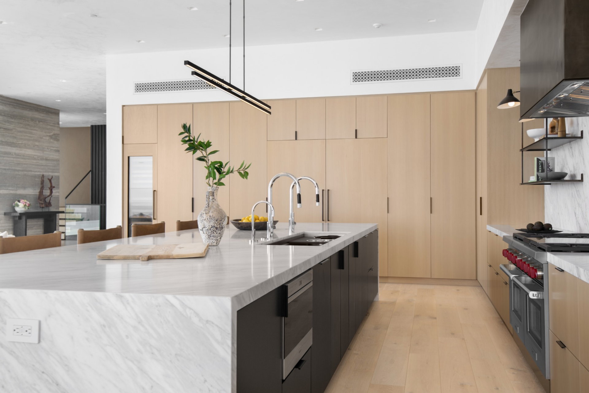 Along the walls, pale wood cabinetry keeps the storage streamlined, while a marble backsplash, wine fridge, and high-end appliances, including a large range beneath a dark metal hood, give the space its more functional edge.