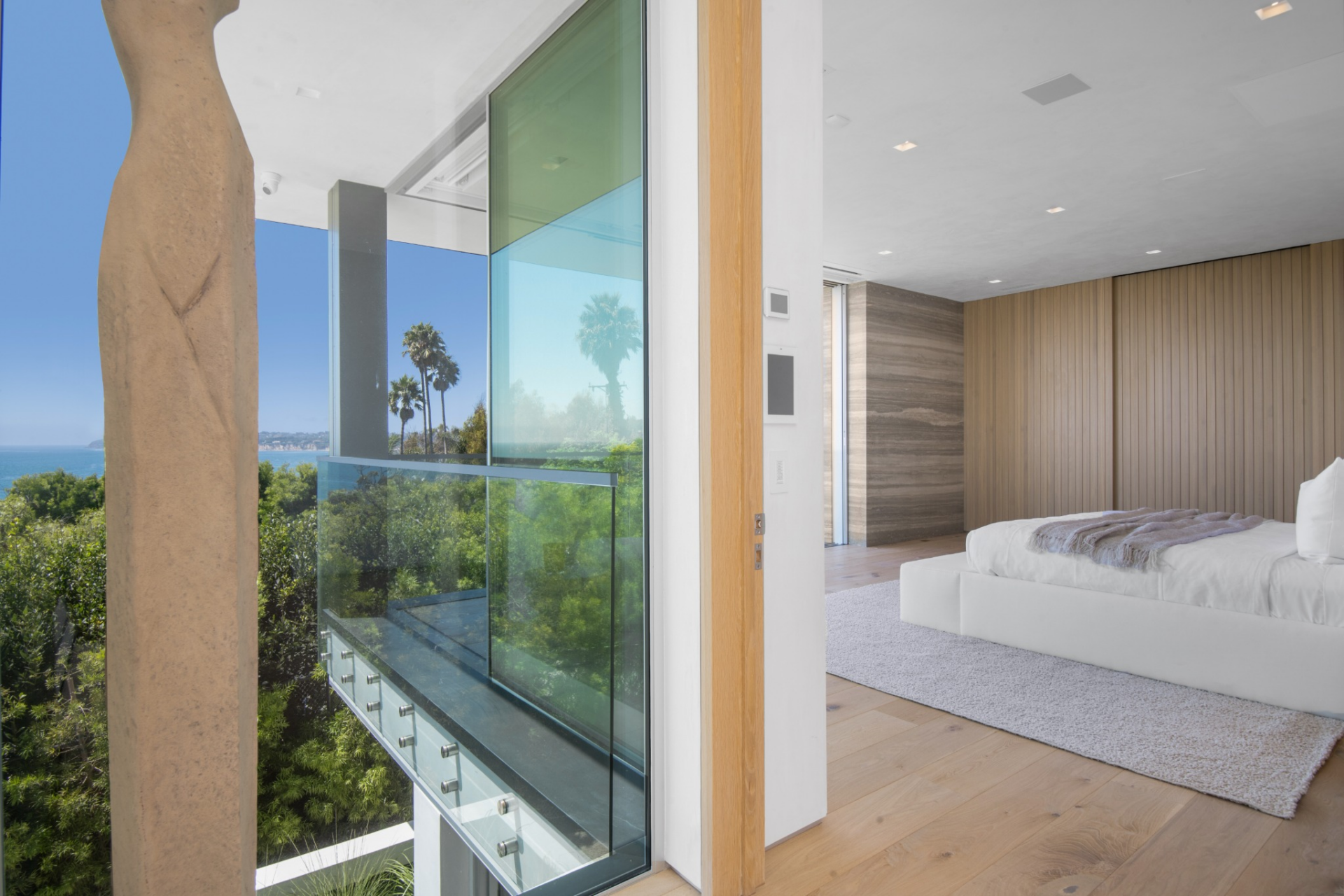 The primary suite continues the home’s clean, open layout with a sleeping area set beside a full-height corner that opens to a glass guardrail overlooking the coastline.