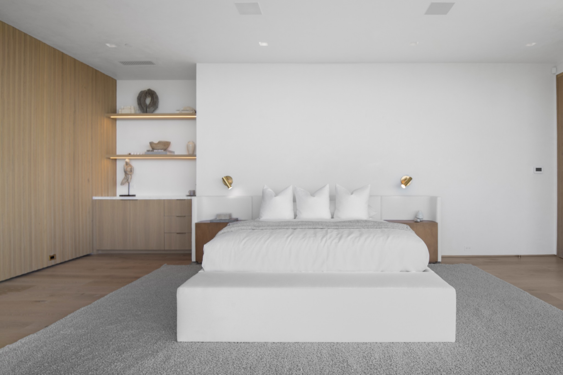 Inside, the room stays restrained, with a low platform bed, wide-plank oak floors, vertical wood paneling, and built-in shelving and storage worked into one side of the space.