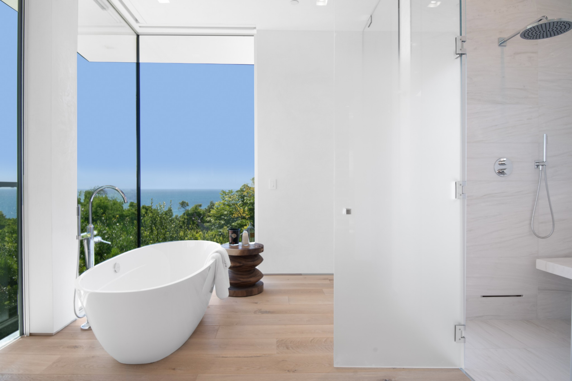 Across from it, a glass-enclosed walk-in shower sits beside a freestanding soaking tub placed right at the window, giving that side of the room a direct view out to the water.