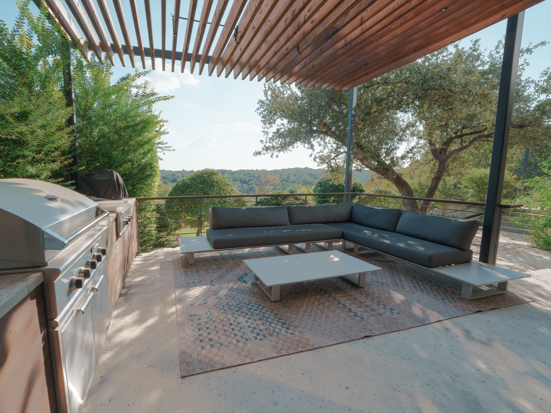 A built-in stainless steel grill sits beside an L-shaped sectional and low-profile tables, forming a dedicated zone for cooking and casual dining against a backdrop of mature trees.