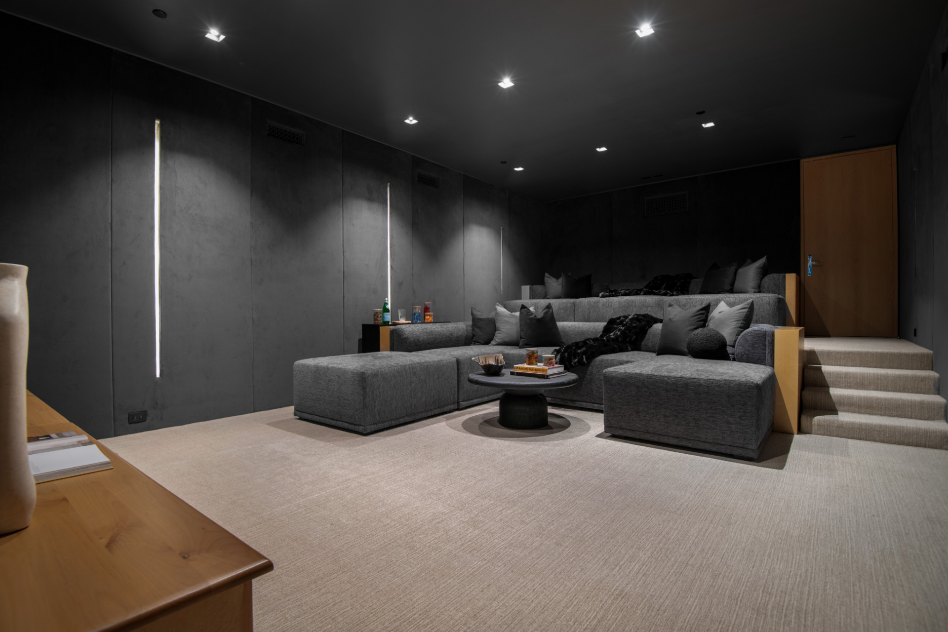 Black walls, a black ceiling, tiered built-in seating, and a large projection screen give the room a more enclosed feel, while slim vertical light strips and low wood detailing break up the darker palette.