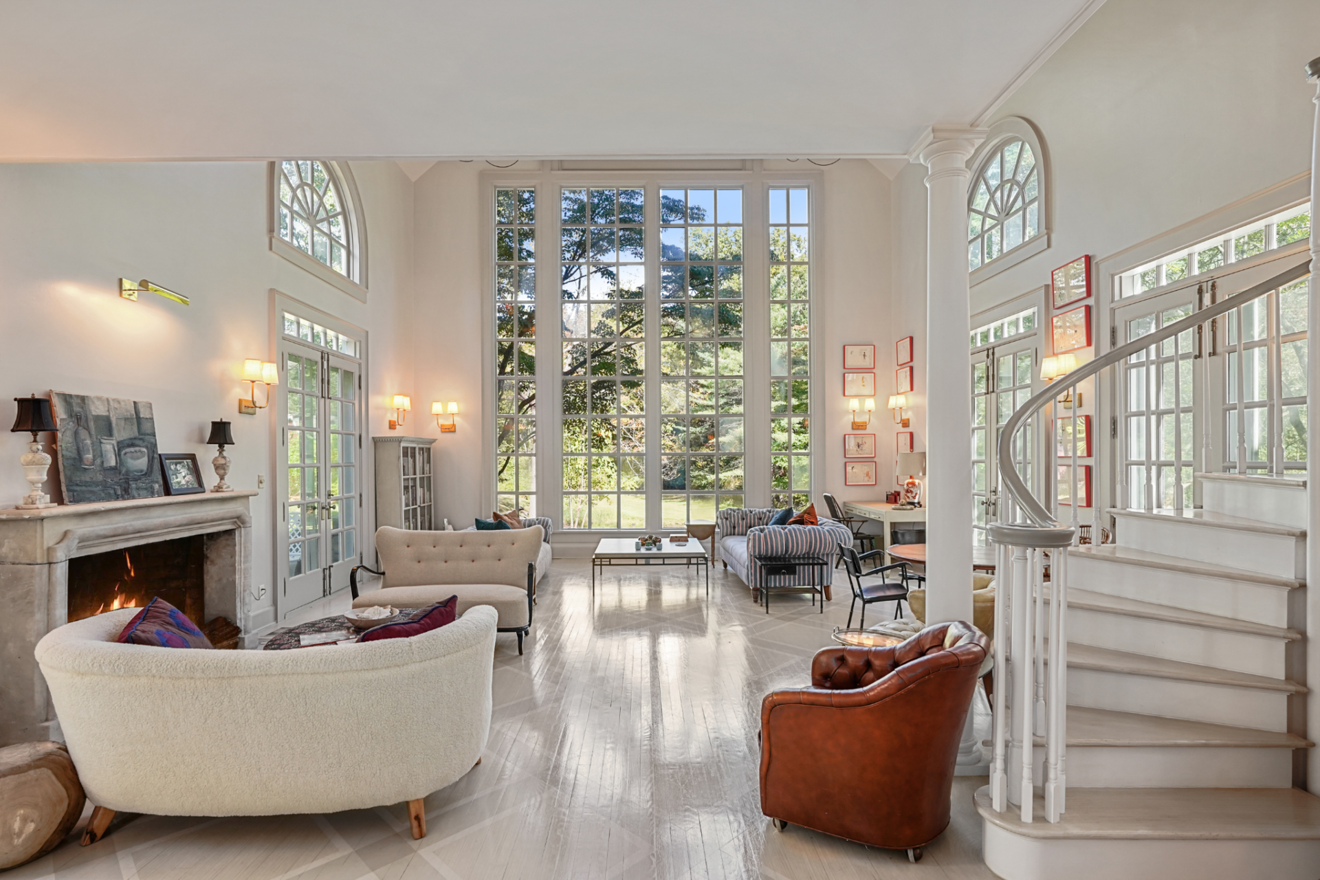 A limestone fireplace anchors one side, while a sweeping staircase and a series of tall, divided-light windows give the space its more formal structure.