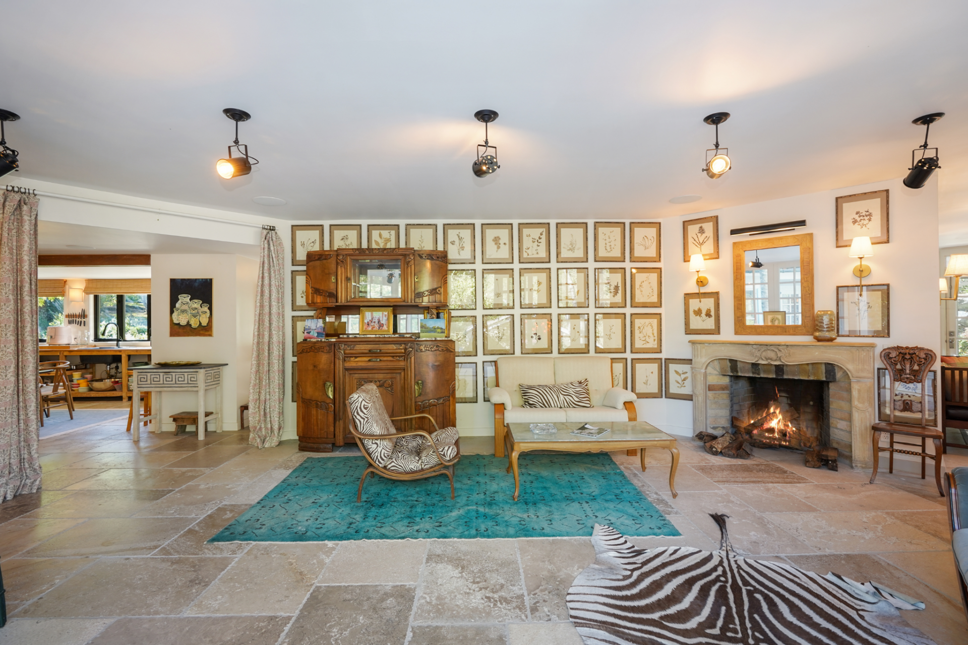 Here, limestone floors replace the pale wood, and a carved stone fireplace sits at one wall beneath a mix of framed artwork and a large mirror. 