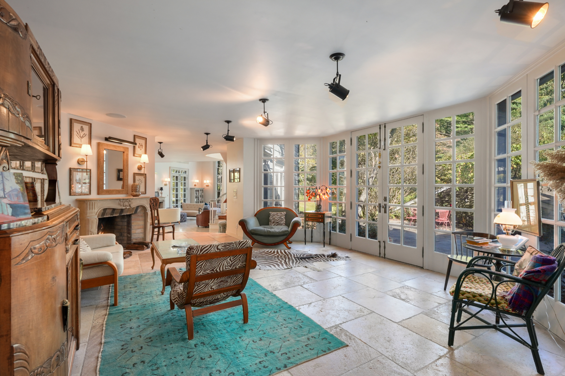 French doors and full-height windows keep the room tied to the grounds, while the furnishings feel more collected, with vintage wood pieces, mixed seating, and patterned accents giving this part of the house a more casual, lived-in setup.
