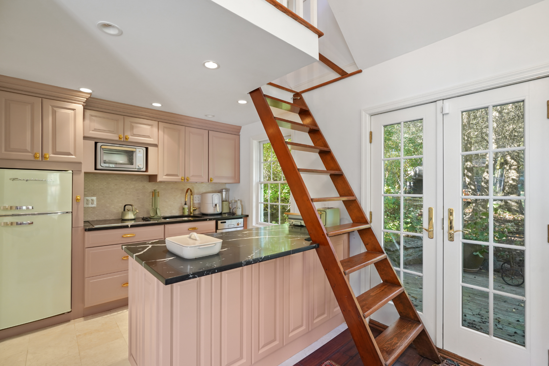 Tucked beneath the loft, the kitchenette features blush-toned cabinetry, dark counters, brass hardware, and a compact island.