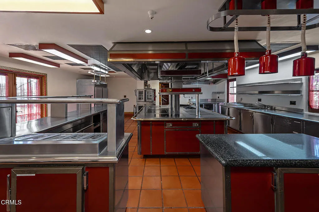 Its layout is centered on production, with multiple stainless prep surfaces, commercial-grade fixtures, and a large central cooking island positioned beneath an oversized ventilation hood.