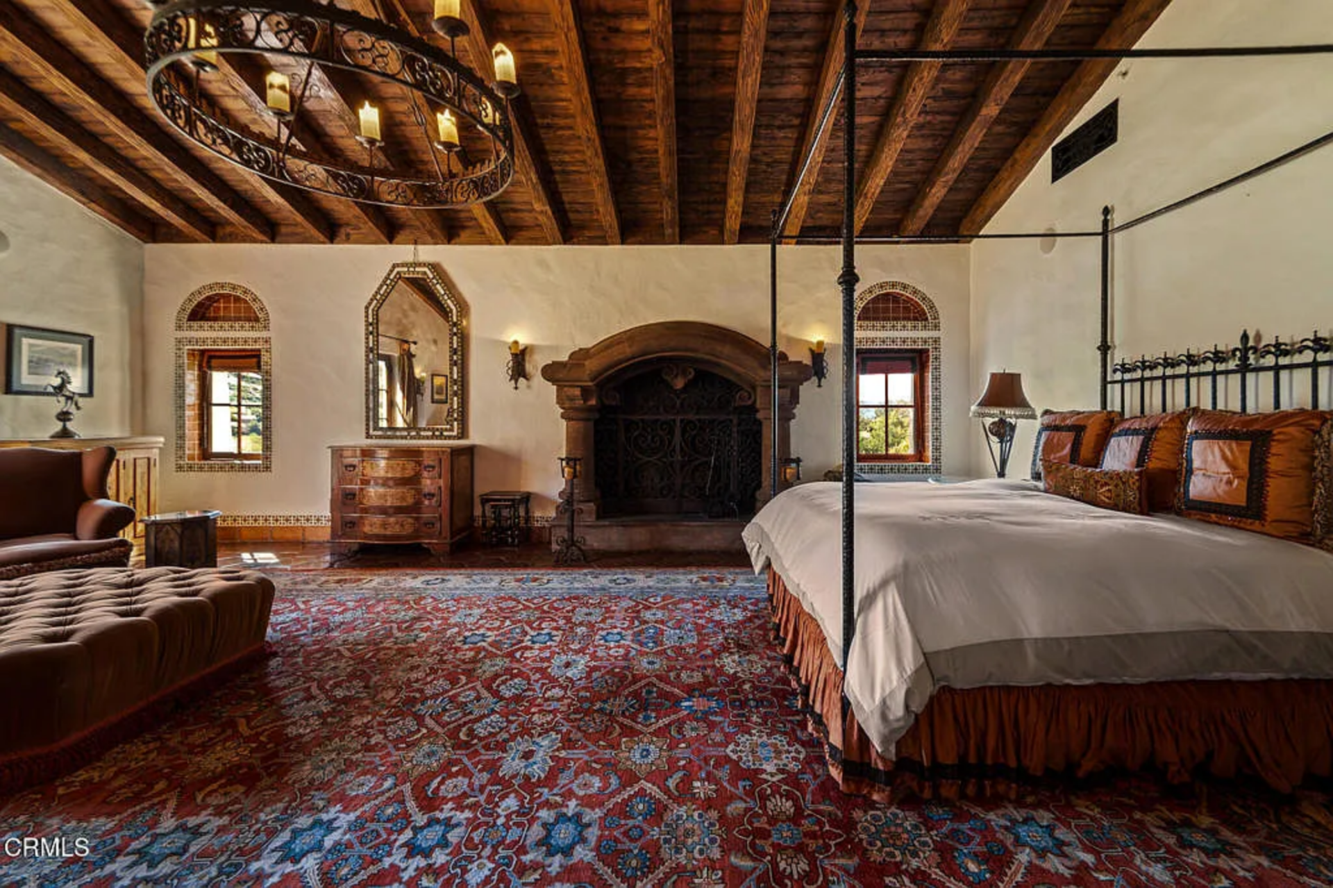A four-poster iron bed sits to one side, while a lounge chair and ottoman create a smaller seating zone closer to the balcony doors, giving the space enough room to function beyond just sleeping.