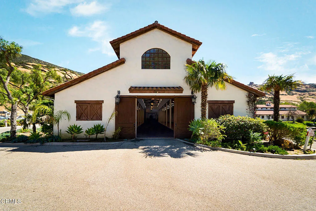 The event barn is set apart from the main villa, with a broad paved courtyard and an exterior finished in white stucco, dark wood doors, and a red tile roof. 