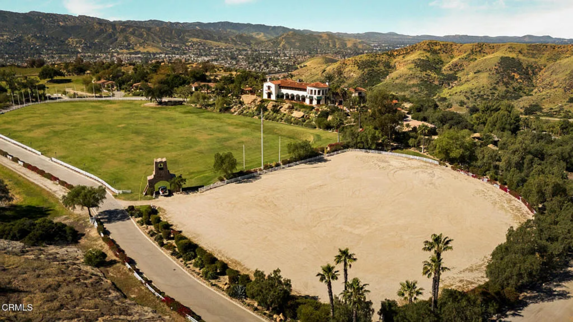 A large riding arena sits below the main villa, bordered by fencing and access roads, while the stable complex is positioned farther into the landscape.