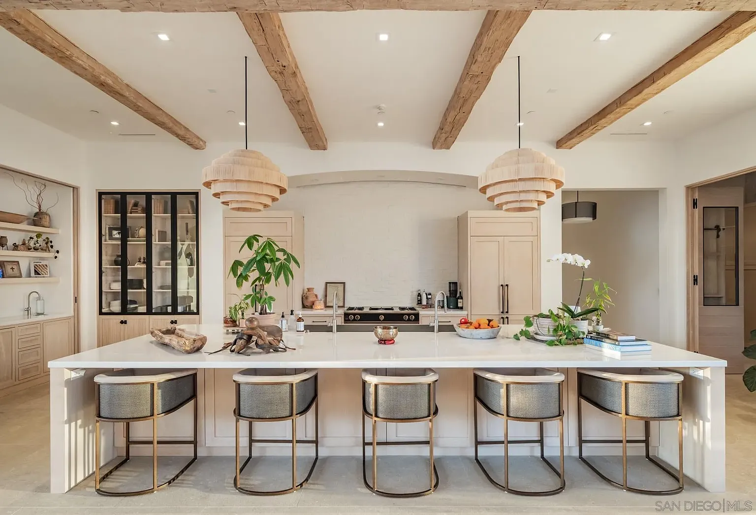 Behind the living area, the kitchen stays fully open to the same shared space, with a long central island placed beneath a pair of layered pendant lights. 