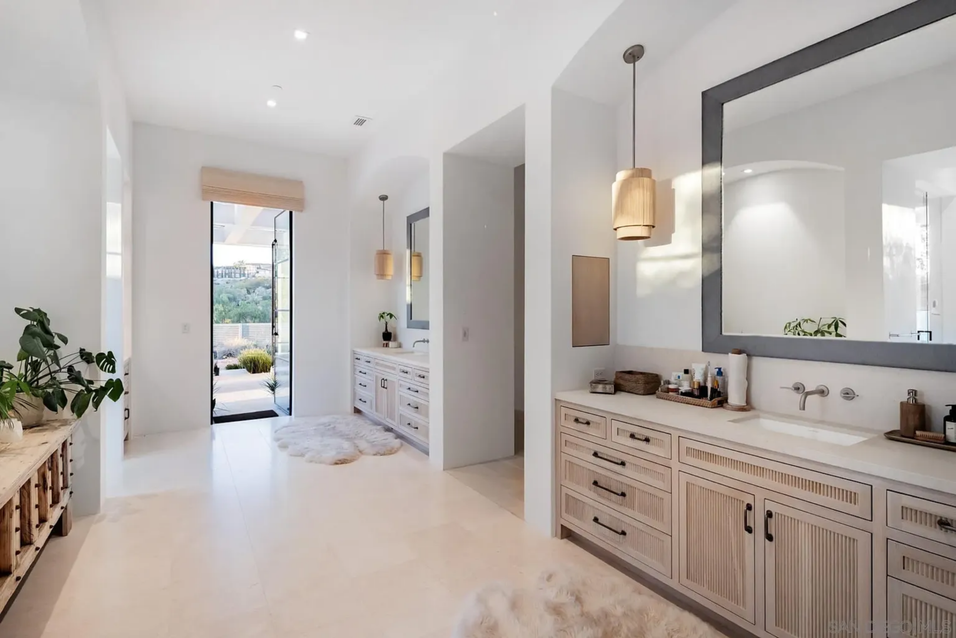 From the bedroom, the bathroom opens into a long, bright space lined with pale cabinetry, oversized mirrors, and hanging pendant lights that bring a softer look to the vanity walls. 