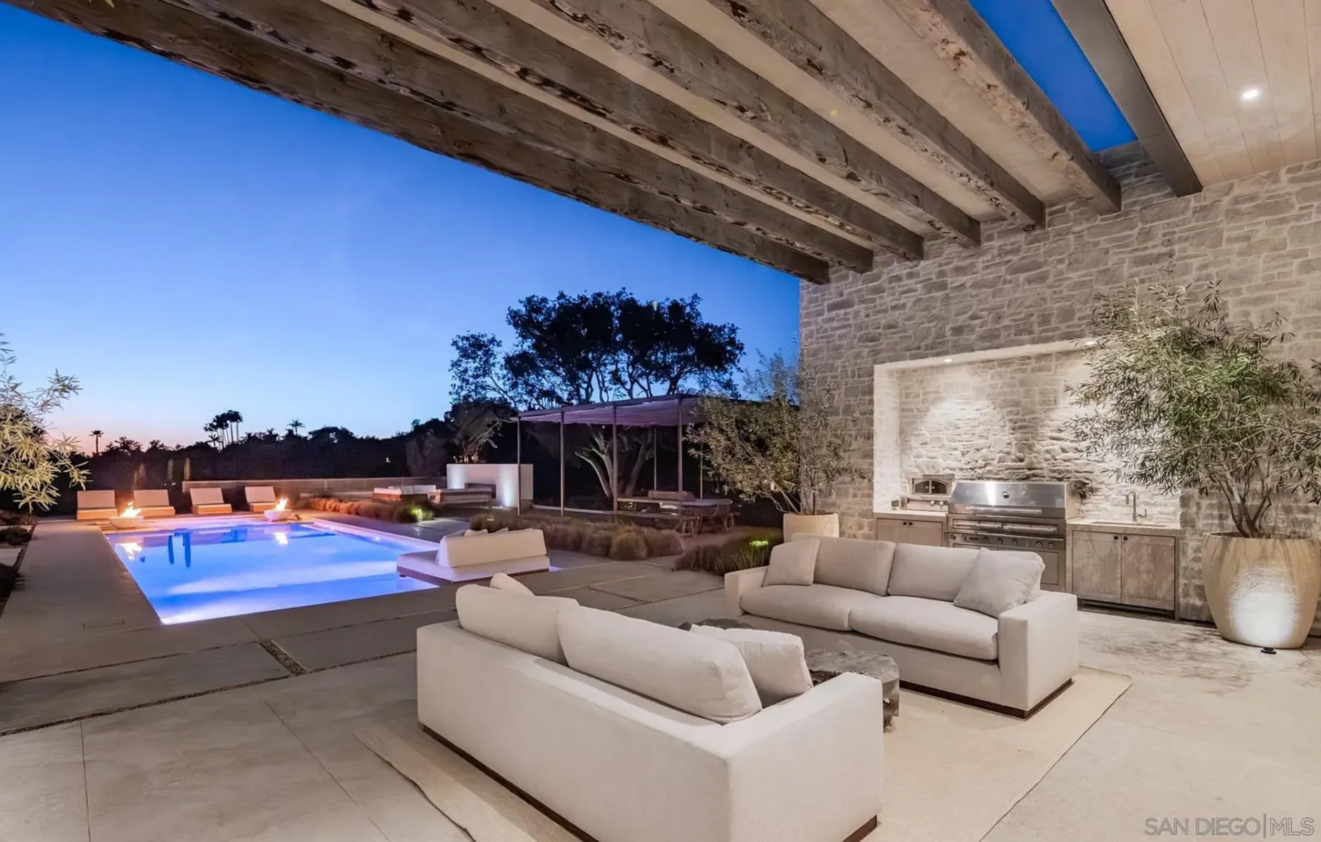The outdoor kitchen lounge is built as an extension of the living spaces, tucked beneath a deep covered terrace with exposed wood beams, stone walls, and a built-in cooking station. 