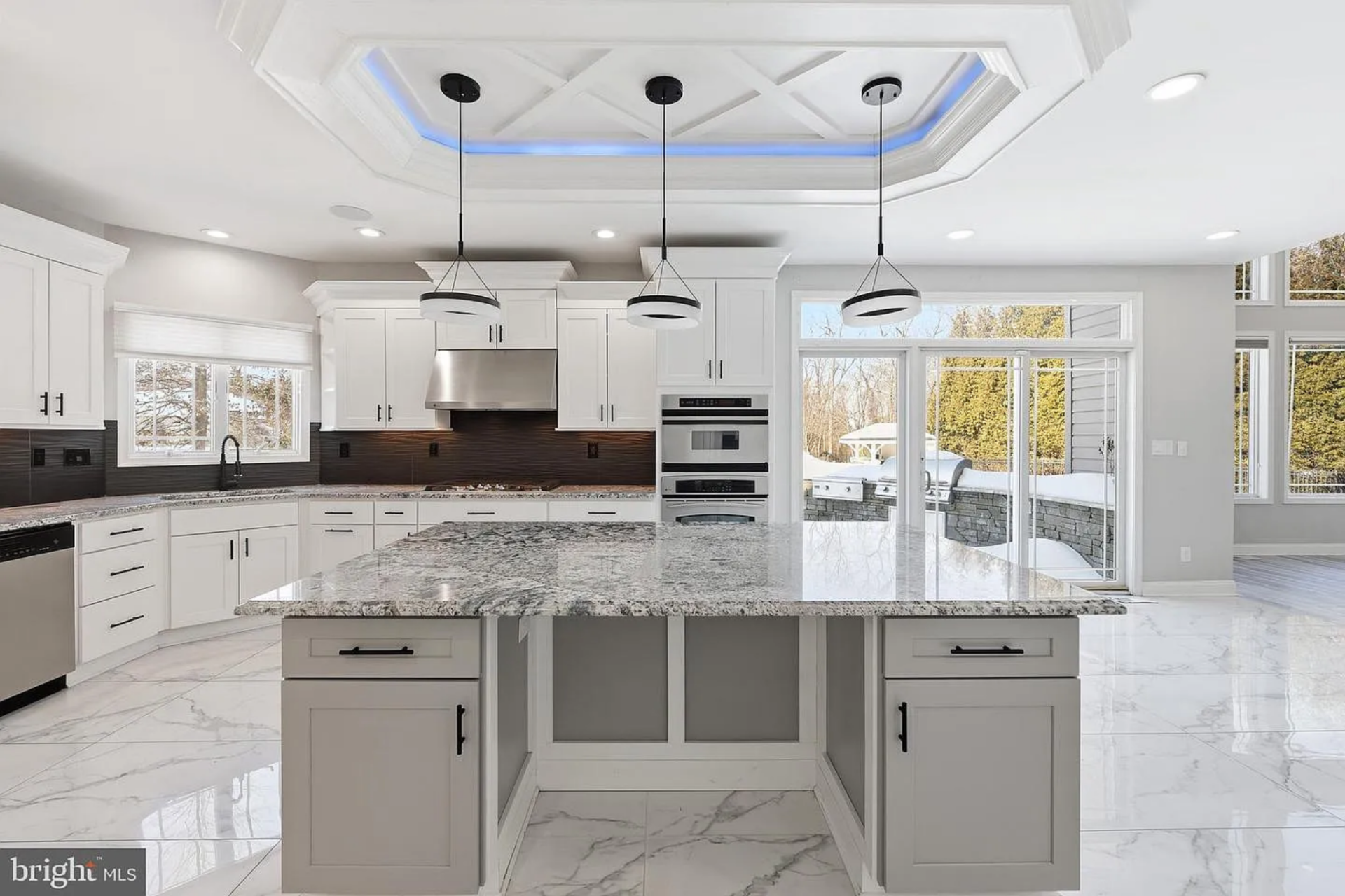 Above, a tray ceiling with detailed millwork and subtle blue accent lighting frames three contemporary ring pendant fixtures, giving the kitchen a defined architectural focal point.