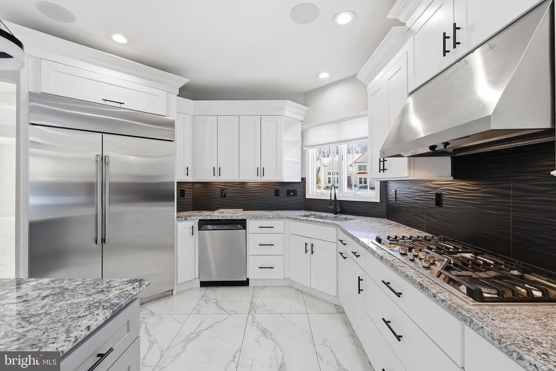 Along one wall, stainless steel appliances, including a professional-grade gas cooktop with a sleek vent hood, double wall ovens, and an oversized refrigerator, line up between custom cabinetry.