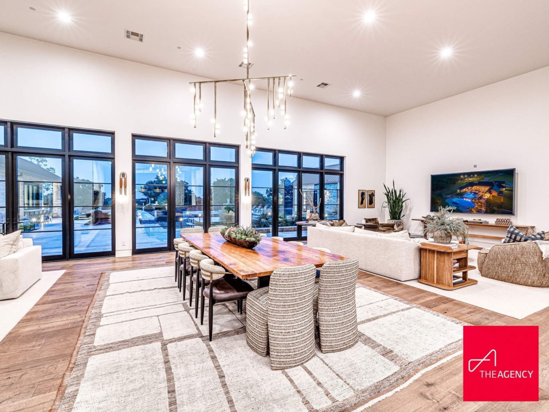 Moving toward the center of the room, the dining area features a long, solid wood table positioned beneath a modern linear chandelier with suspended glass elements.