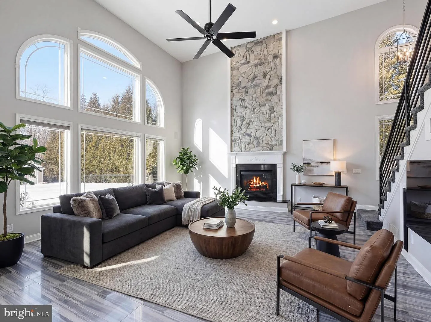 A large sectional and accent chairs are arranged around a central coffee table, creating a comfortable seating area beneath a modern ceiling fan, while the open staircase along the side reinforces the room’s connection to the upper level.