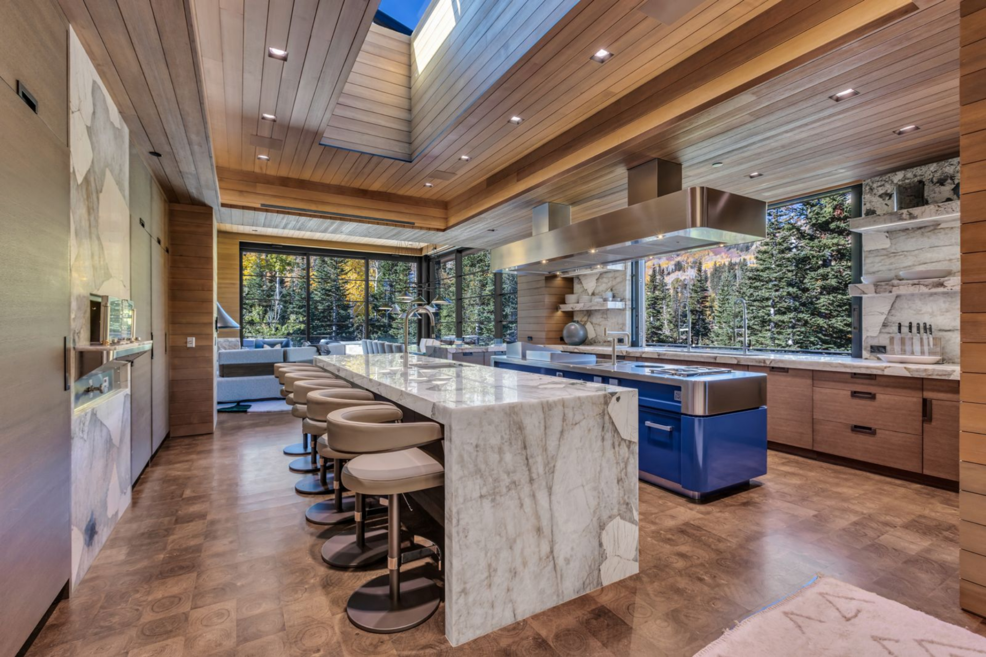 Opposite it, a bold blue range creates a secondary island beneath a stainless-steel hood that floats overhead, its clean lines contrasting with the organic textures of wood and stone.
