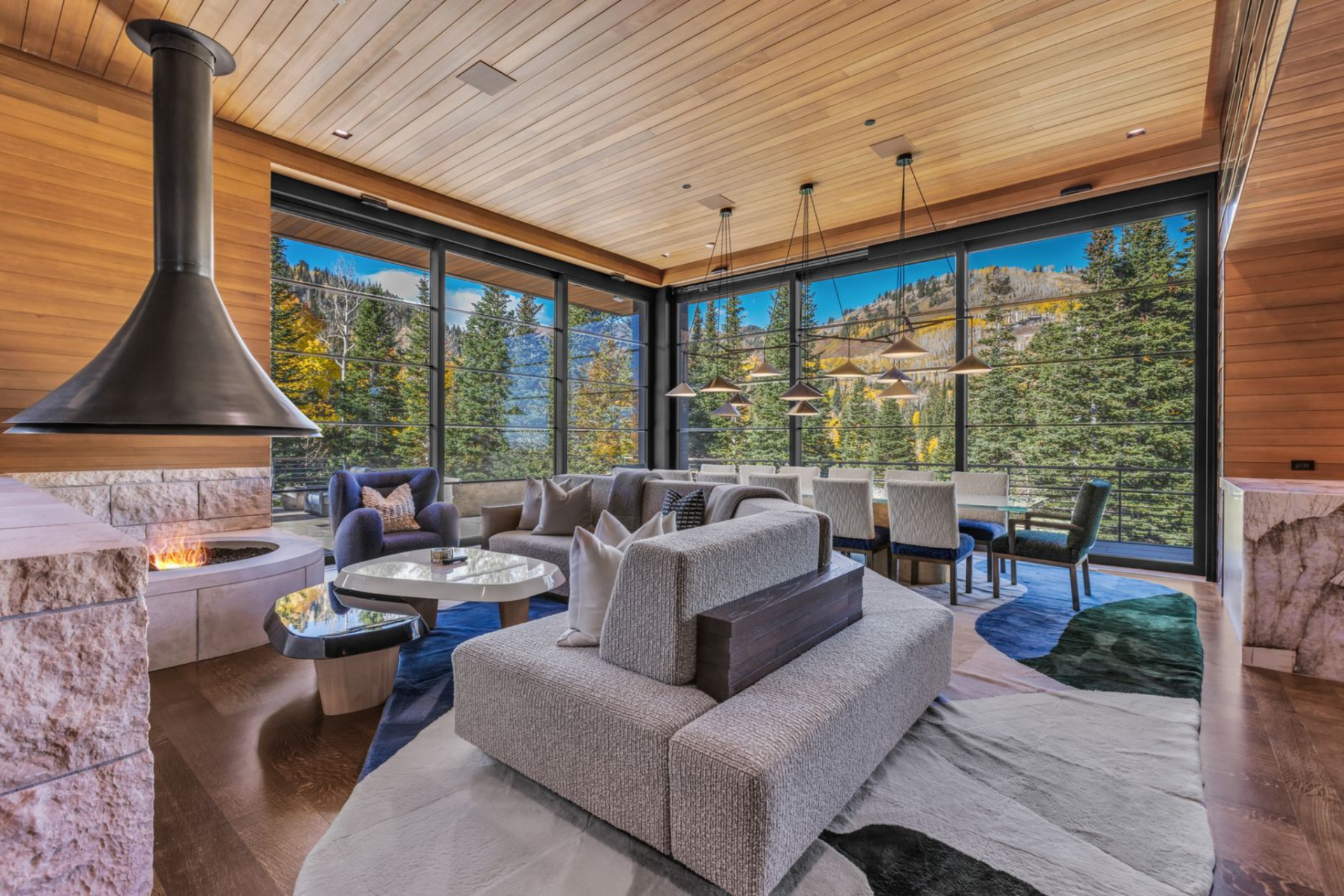 A suspended black metal hood drops from the ceiling above the fire feature, while low-profile sofas and lounge chairs are arranged on a patterned area rug, facing both toward the flames and outward toward the windows.