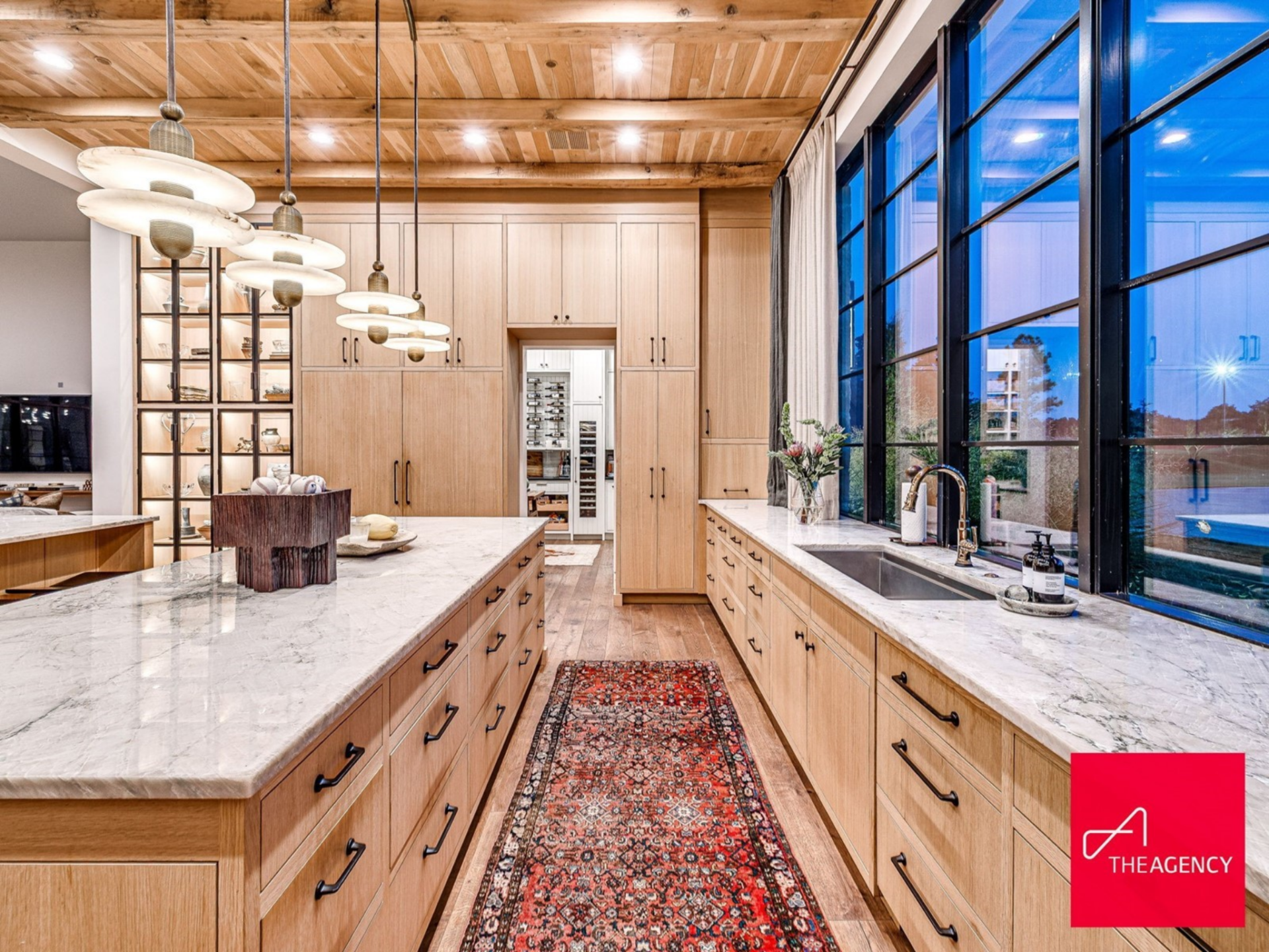 Open shelving frames the cooking alcove, styled with ceramics and functional pieces, while expansive countertops stretch beneath oversized black-framed windows that look out toward the grounds.