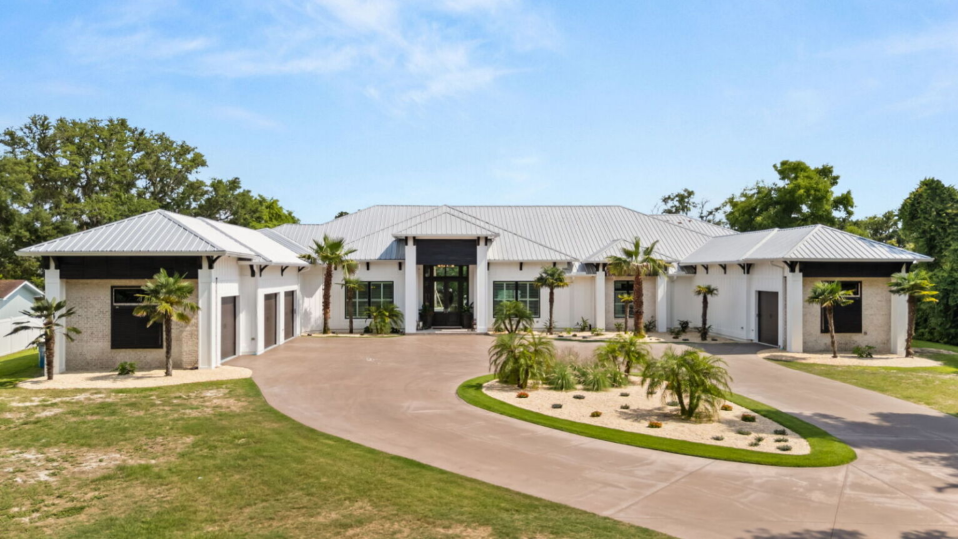 A sweeping circular driveway wraps around a landscaped center island filled with palms and greenery, guiding visitors toward the main entrance while emphasizing the home’s expansive lot.