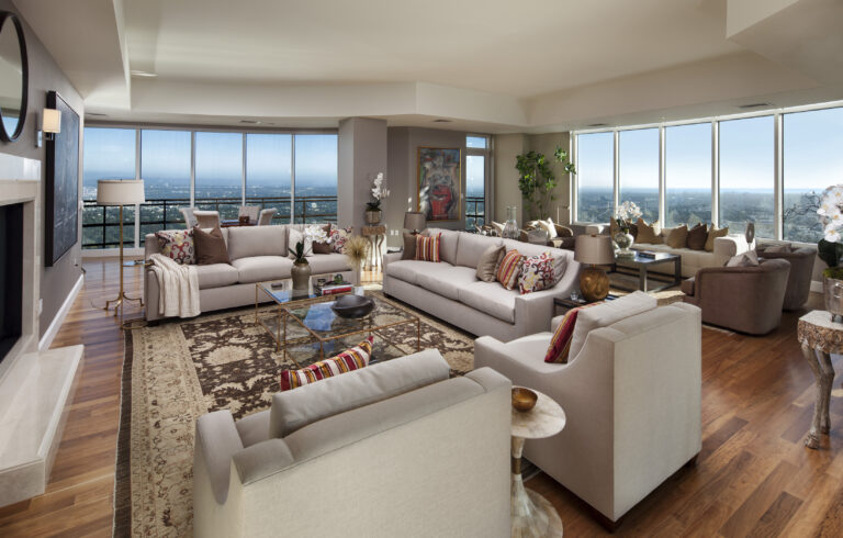 Great room where the seating area is arranged for conversation, with multiple sofas and chairs gathered around a large rug and glass coffee table, while the fireplace adds another warm point of light along the side wall.