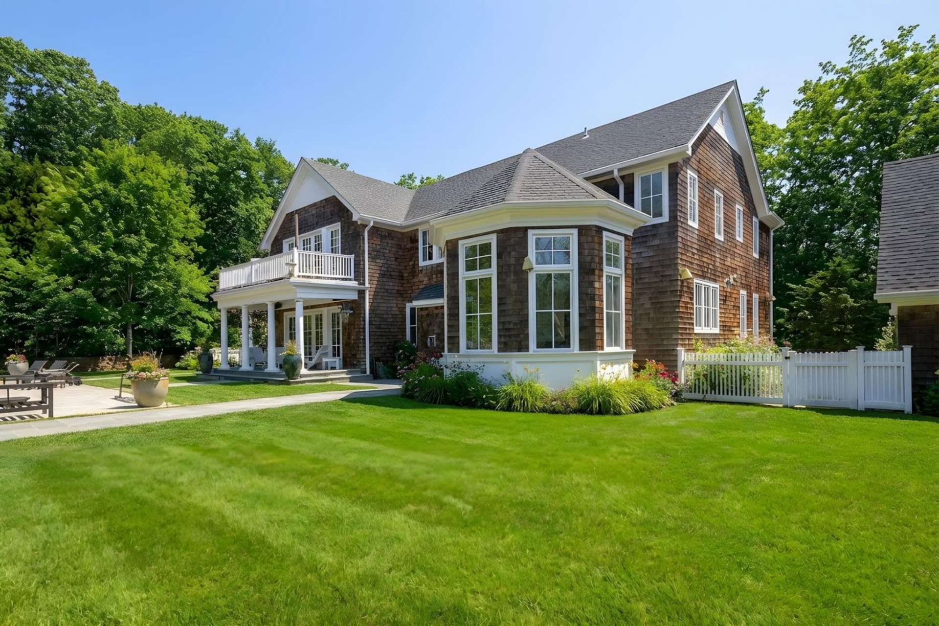 From this angle, the shingle-style exterior, bay window, covered porch, and second-floor balcony all look out over a wide expanse of trimmed grass bordered by trees and low plants.
