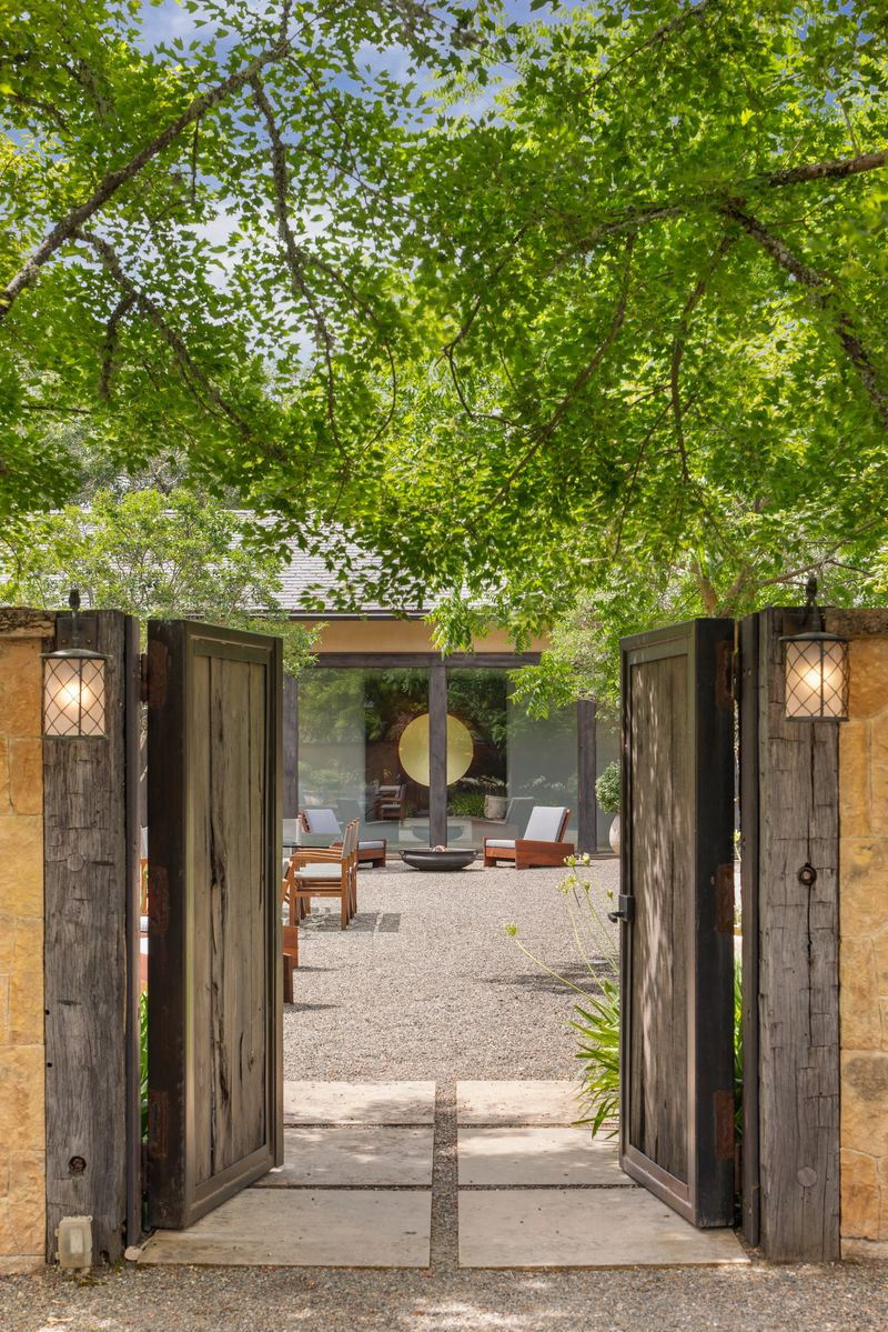 Heading into the Tuscan-inspired main residence, the house opens from a gravel courtyard framed by towering trees, low plants, and broad barn-style wood doors that pull the exterior directly into the entry. 