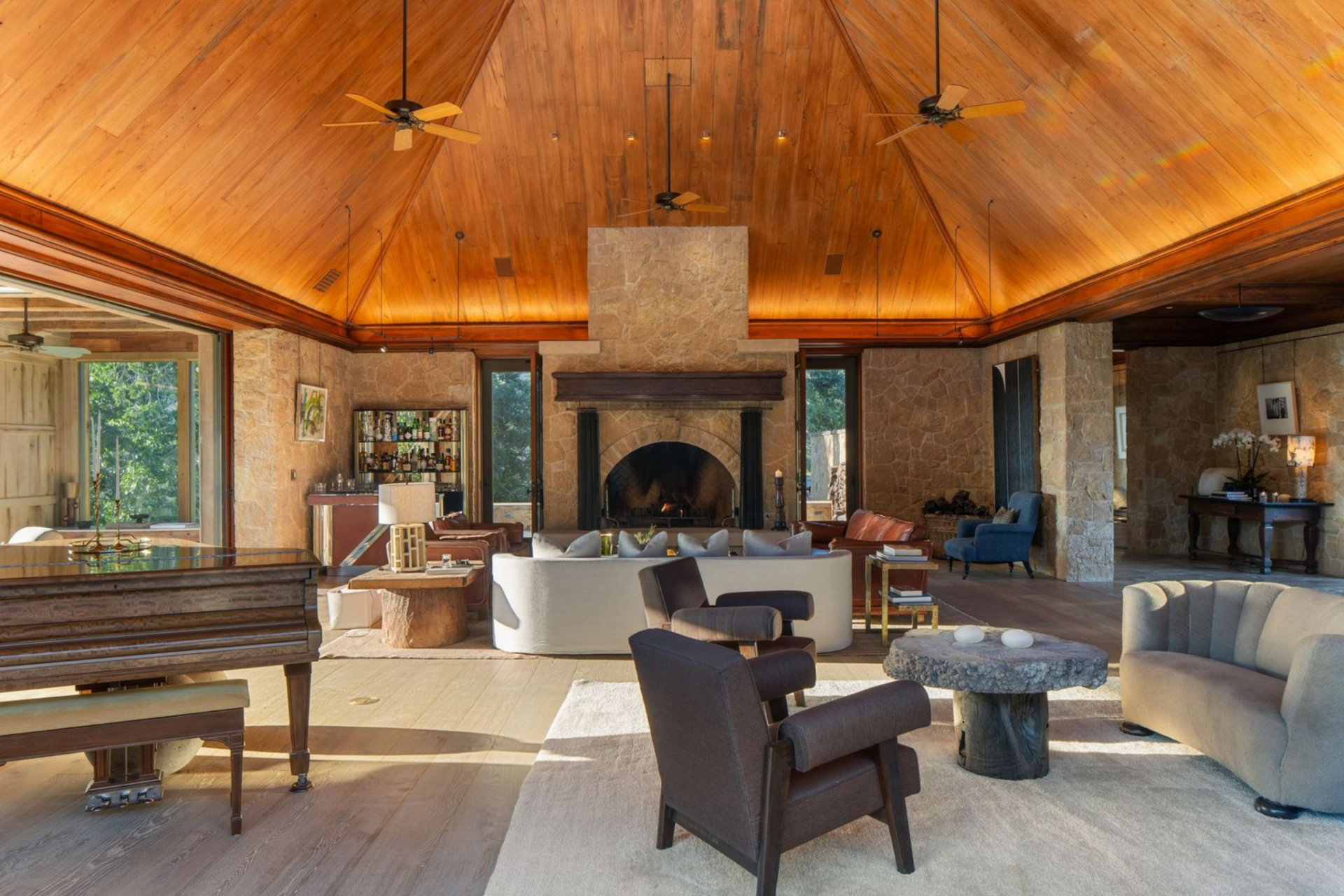 A soaring wood ceiling rises over multiple seating areas, while a massive stone fireplace, grand piano, and built-in bar spread the room across a broad open footprint.