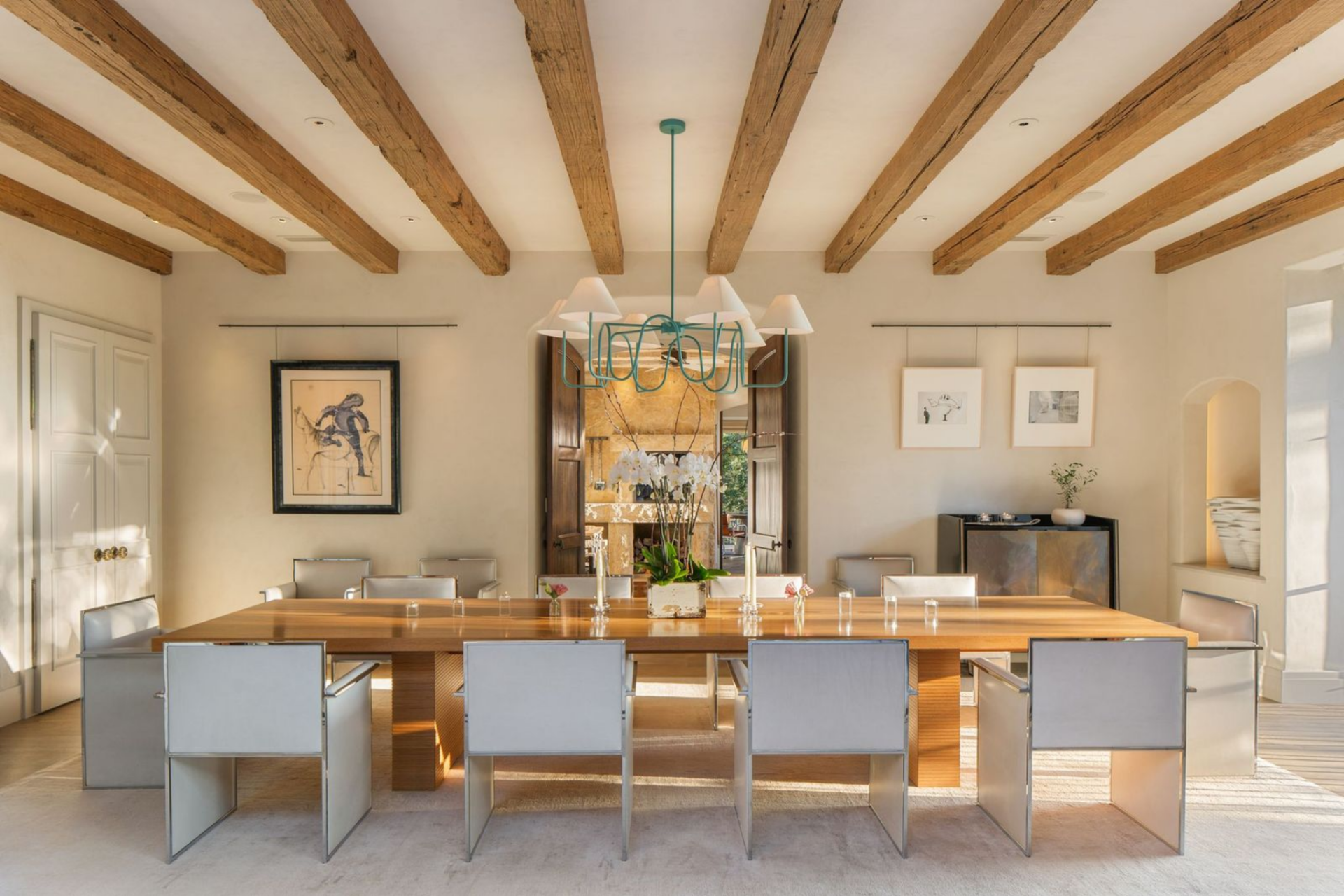Inside, reclaimed wood beams continue overhead, while a long wood dining table sits beneath a sculptural light fixture and is surrounded by a clean lineup of metal chairs.