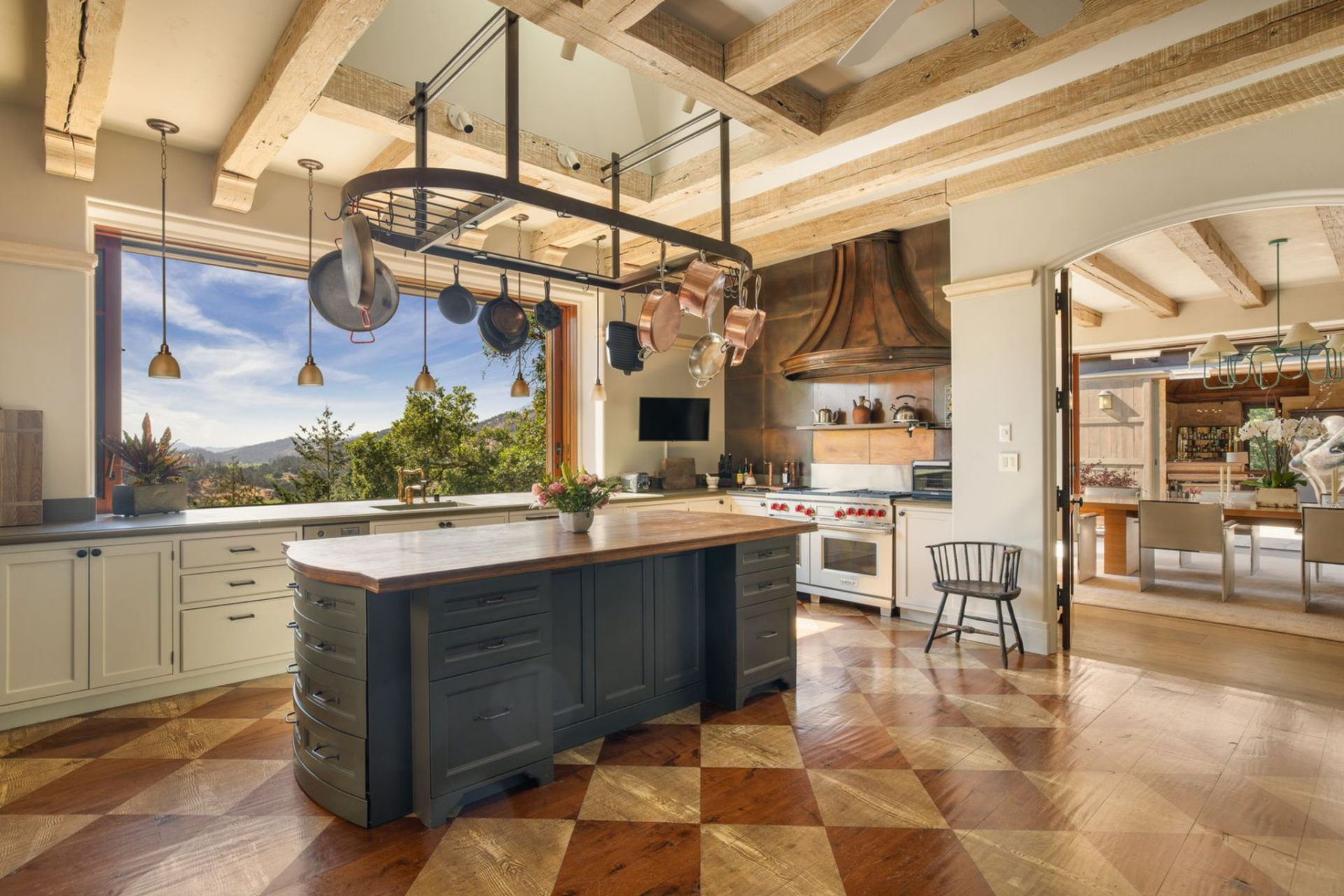 Just beyond the dining room, the kitchen has a patterned wood floor that grounds a large central island with a curved edge, sitting beneath a suspended metal rack of copper and cast-iron cookware.