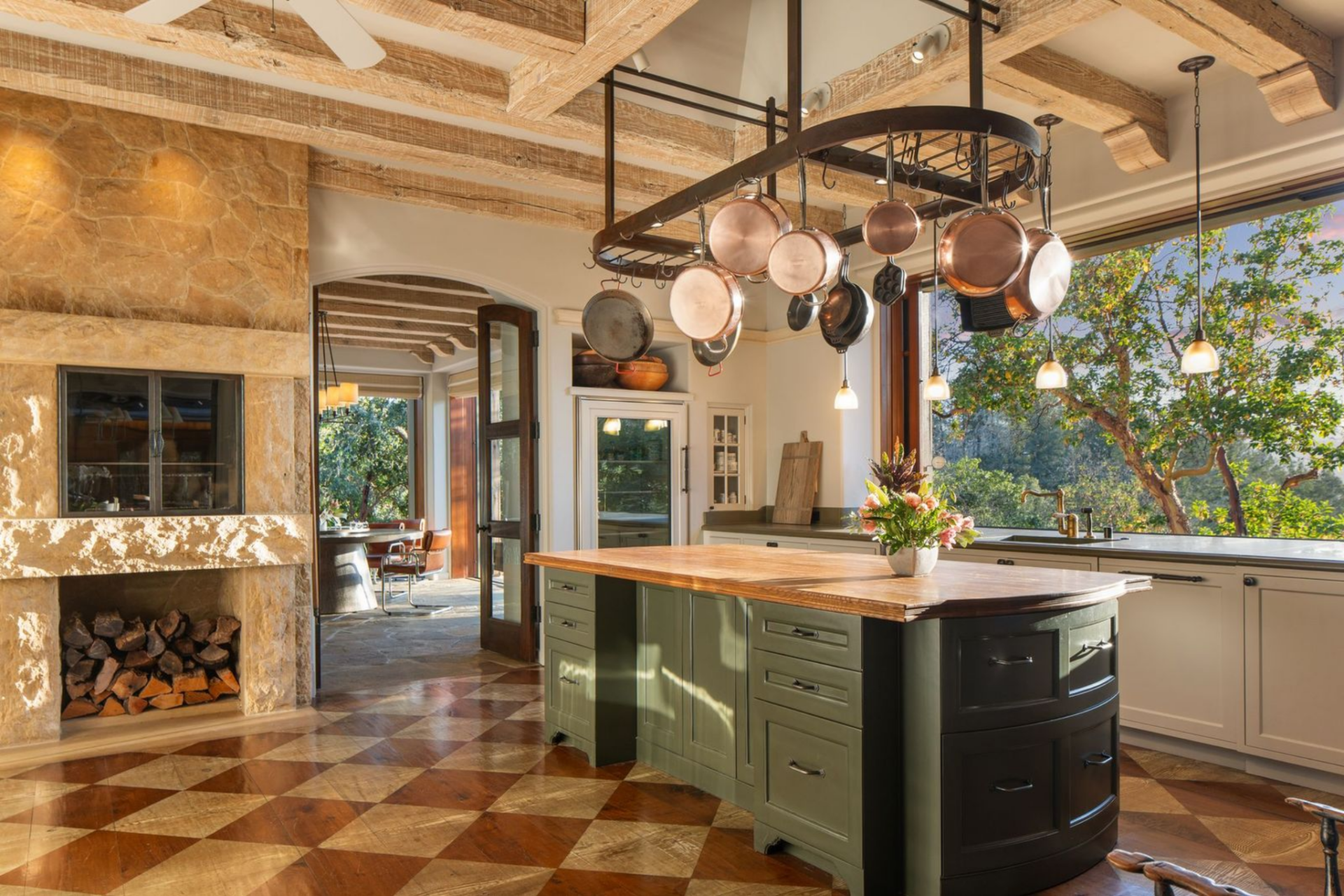 Along the back wall, a custom range is set into a stone surround with a sculpted hood, while the sink is placed beneath a wide window that pulls in light and frames the landscape just beyond.