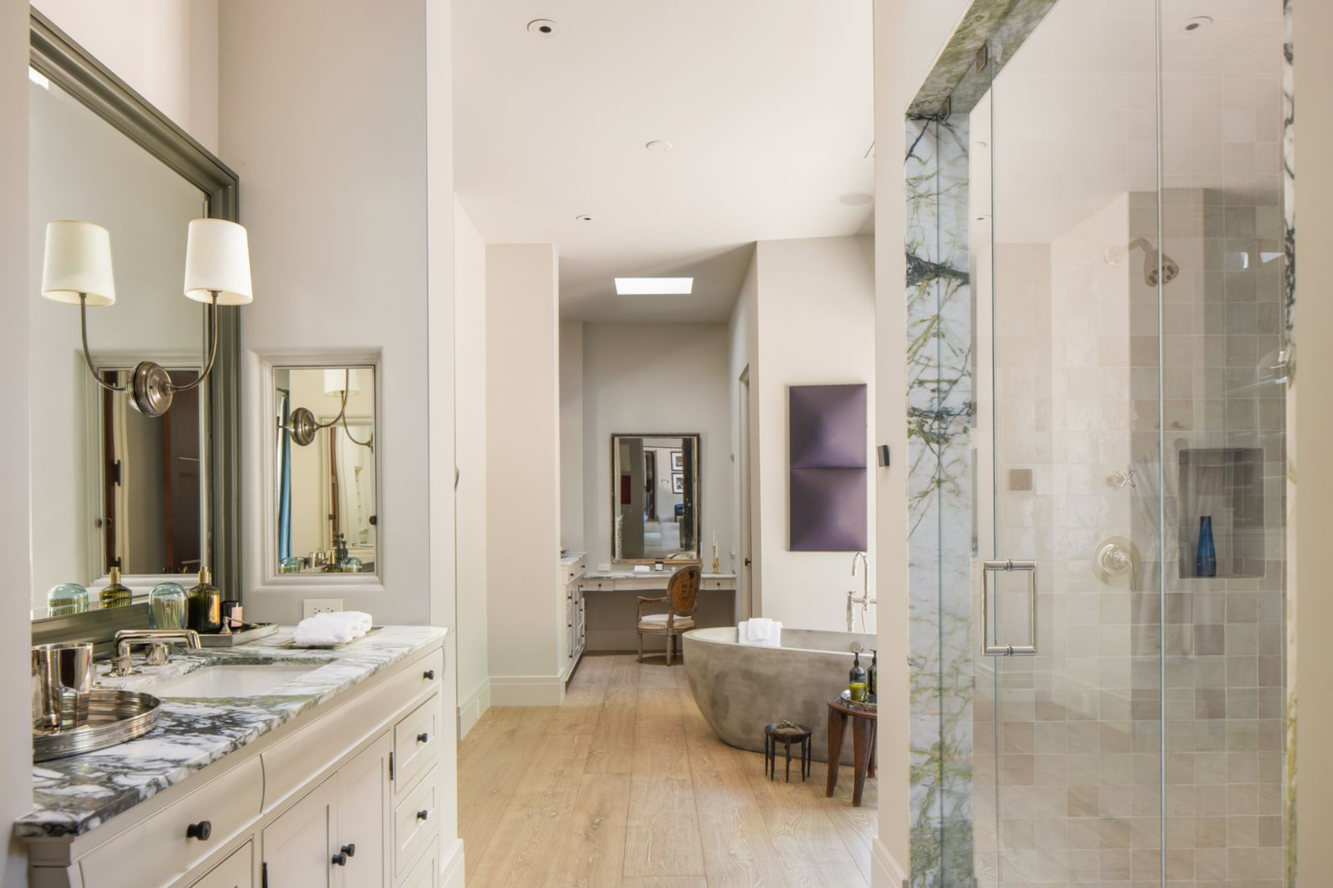 Marble surfaces run along the vanity, the shower is enclosed in glass with veined stone trim, and the freestanding tub is placed in the center.