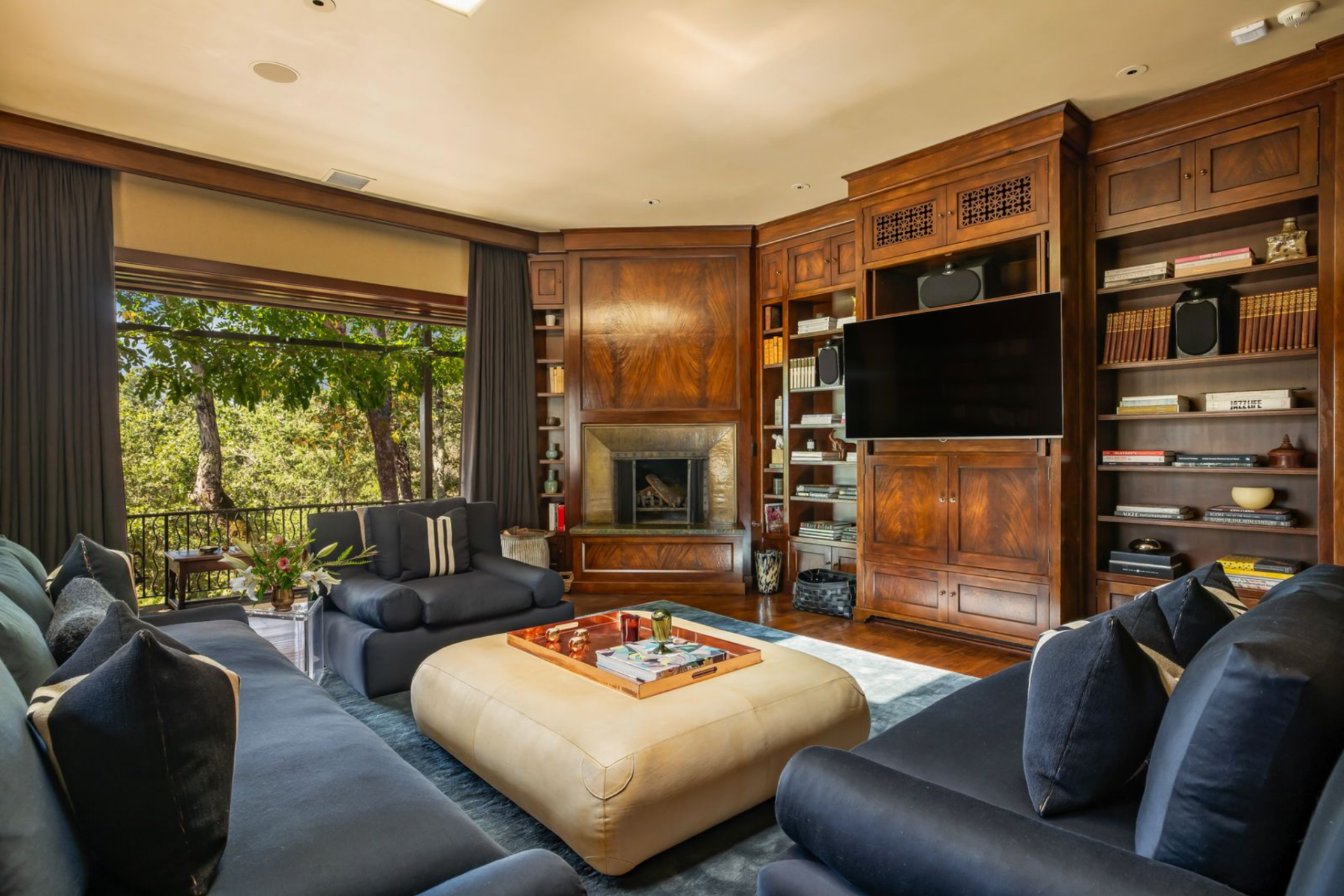 One wall centers on a fireplace with a television mounted within the cabinetry beside it, while seating is gathered tightly around a large central ottoman, with a sofa and matching lounge chairs arranged to face inward.