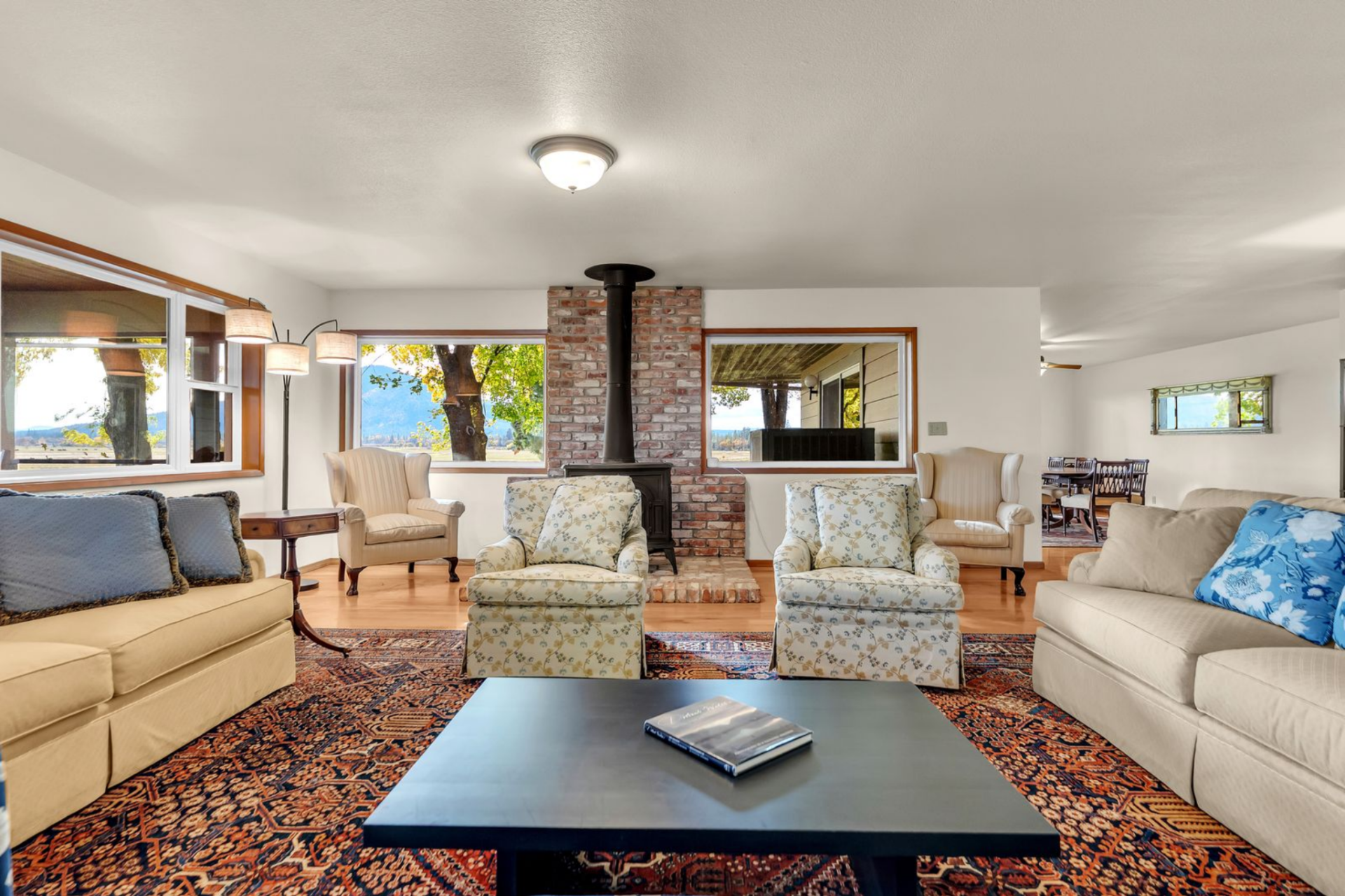 Windows on either side pull in views of the landscape, while the seating area is laid out across a large patterned rug with a mix of sofas, floral armchairs, and wingback chairs gathered around a low square coffee table.