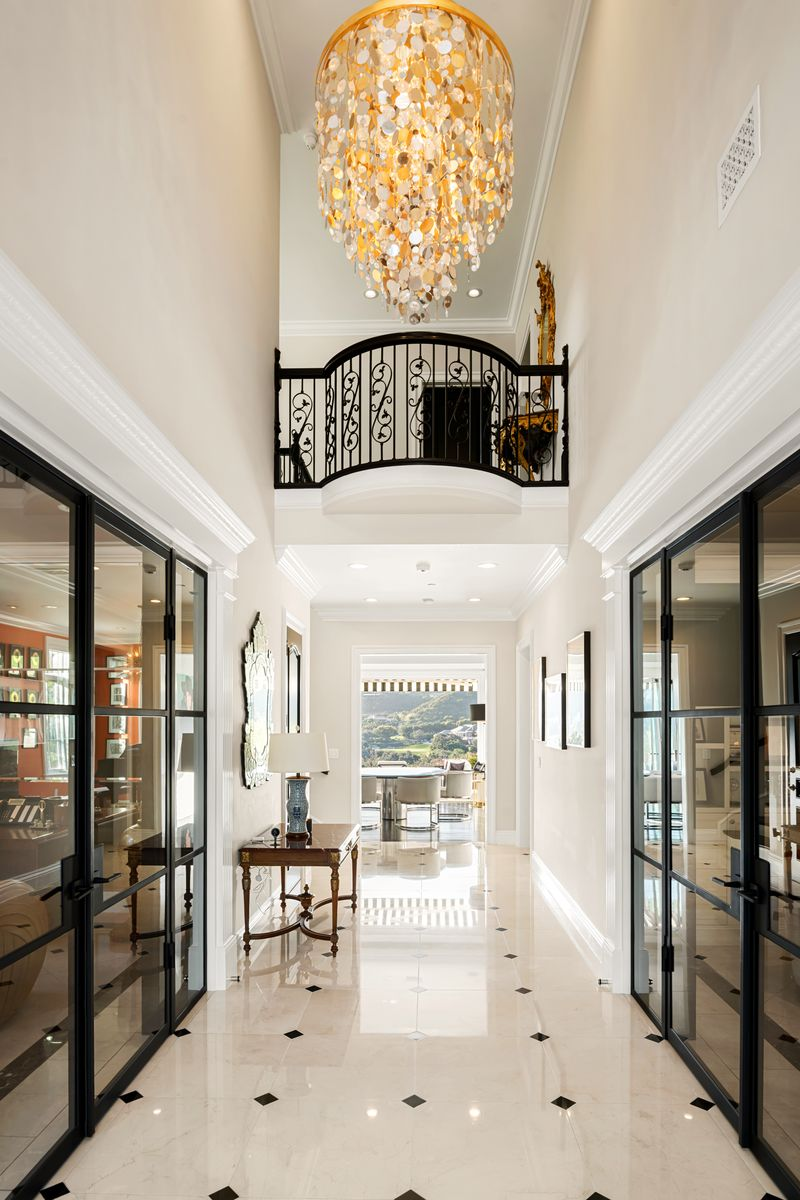 Overhead, a cascading chandelier drops from the double-height ceiling, while a curved balcony with wrought-iron detailing overlooks the entry from above.