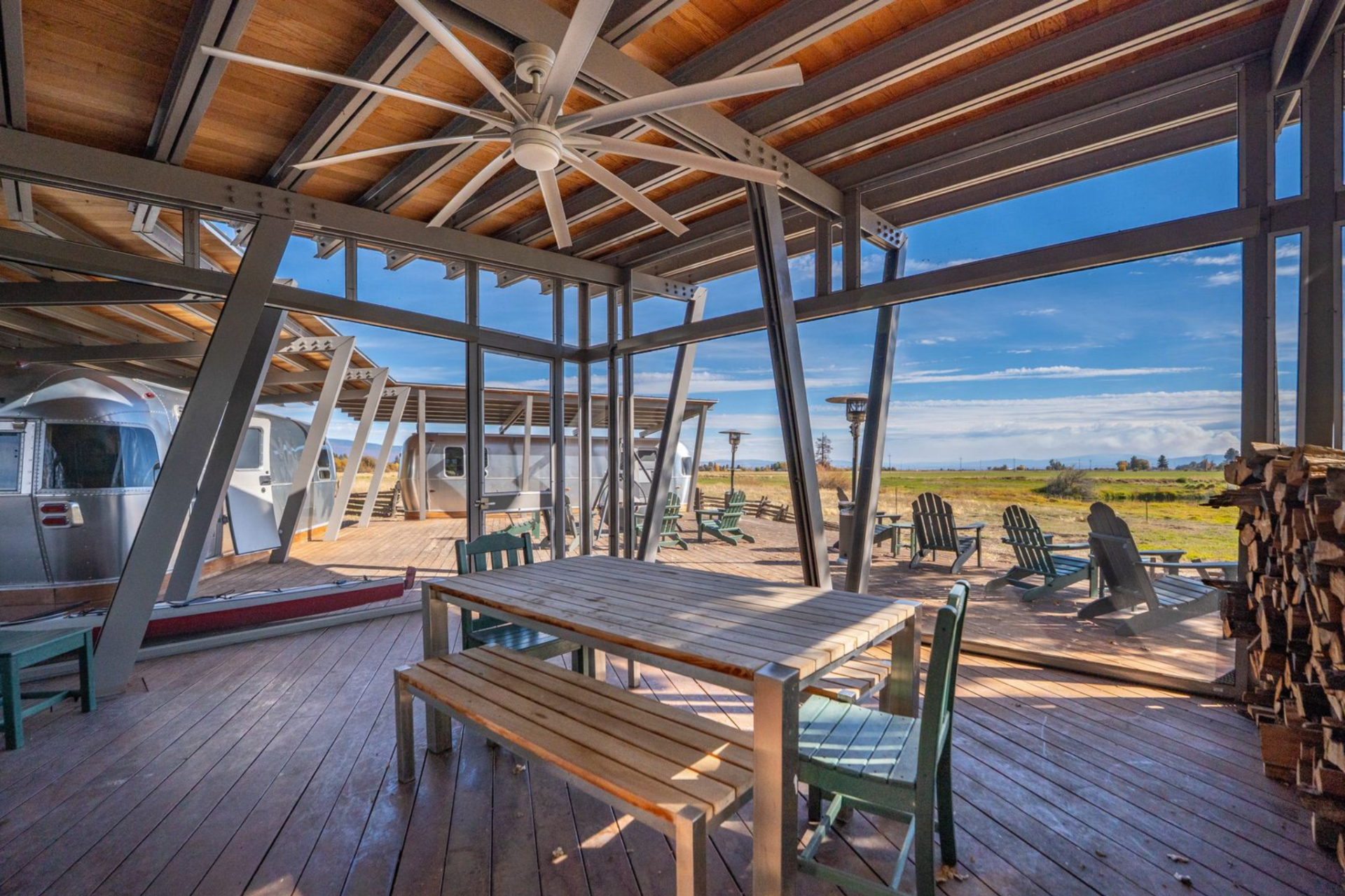 Steel framing and broad glass walls keep the structure open to the landscape, while the wood decking, exposed beams, and long communal table give it the feel of an outdoor lodge rather than a standard camp setup.