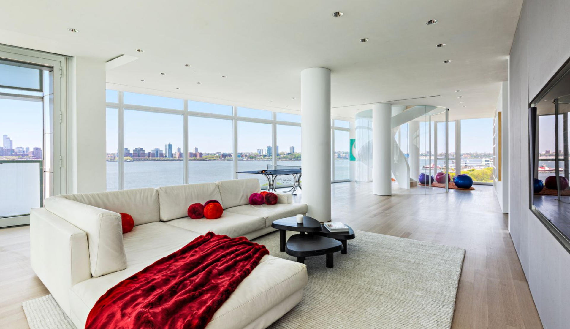 At the center, the main living area is arranged around a low sectional sofa set on a textured rug, with a pair of rounded tables placed in front to keep the seating area grounded without interrupting the openness of the room. 