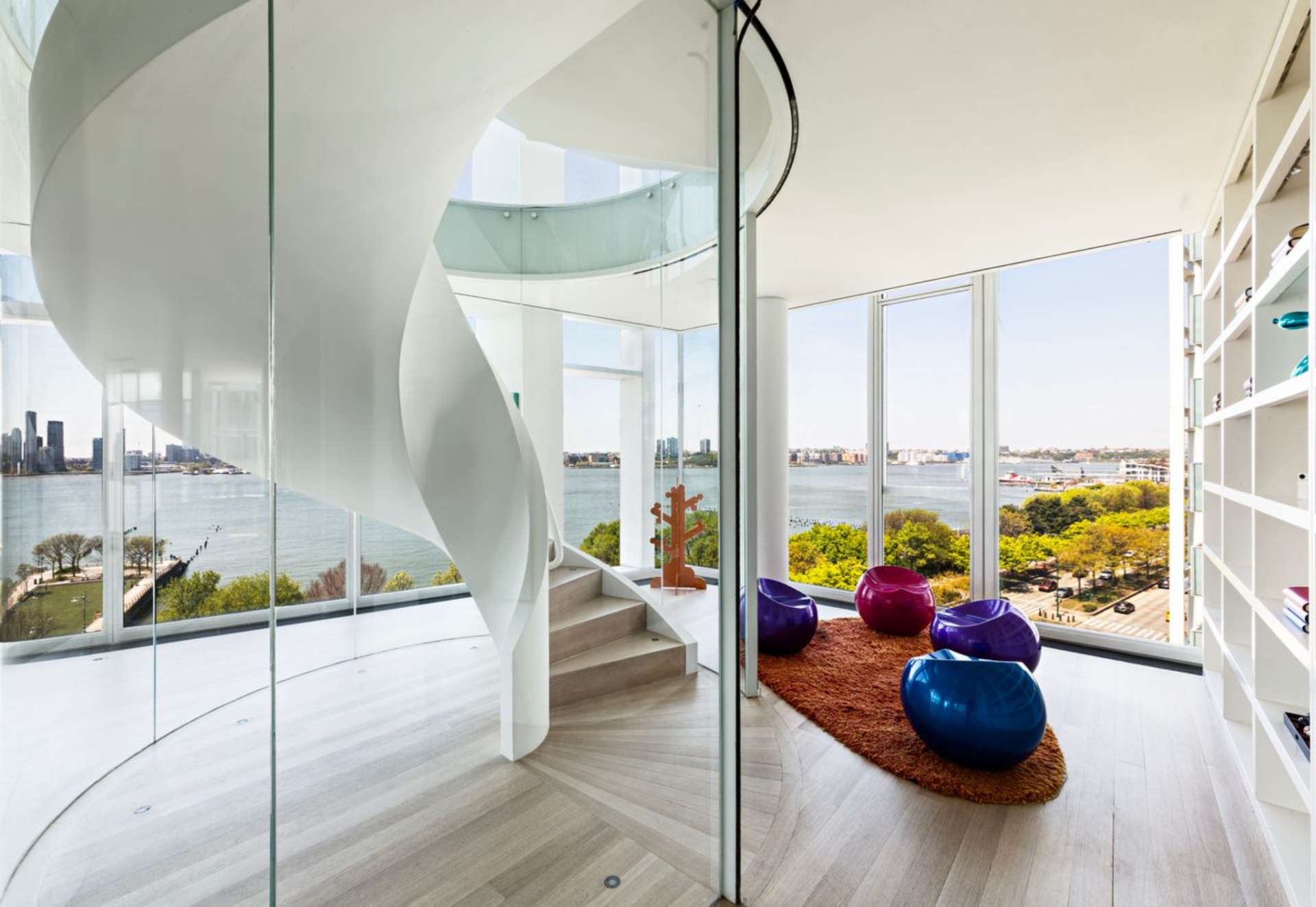 Off to the side, a glass-enclosed spiral staircase that connects all three levels becomes a sculptural focal point, its curved form rising beside a small cluster of colorful seating.