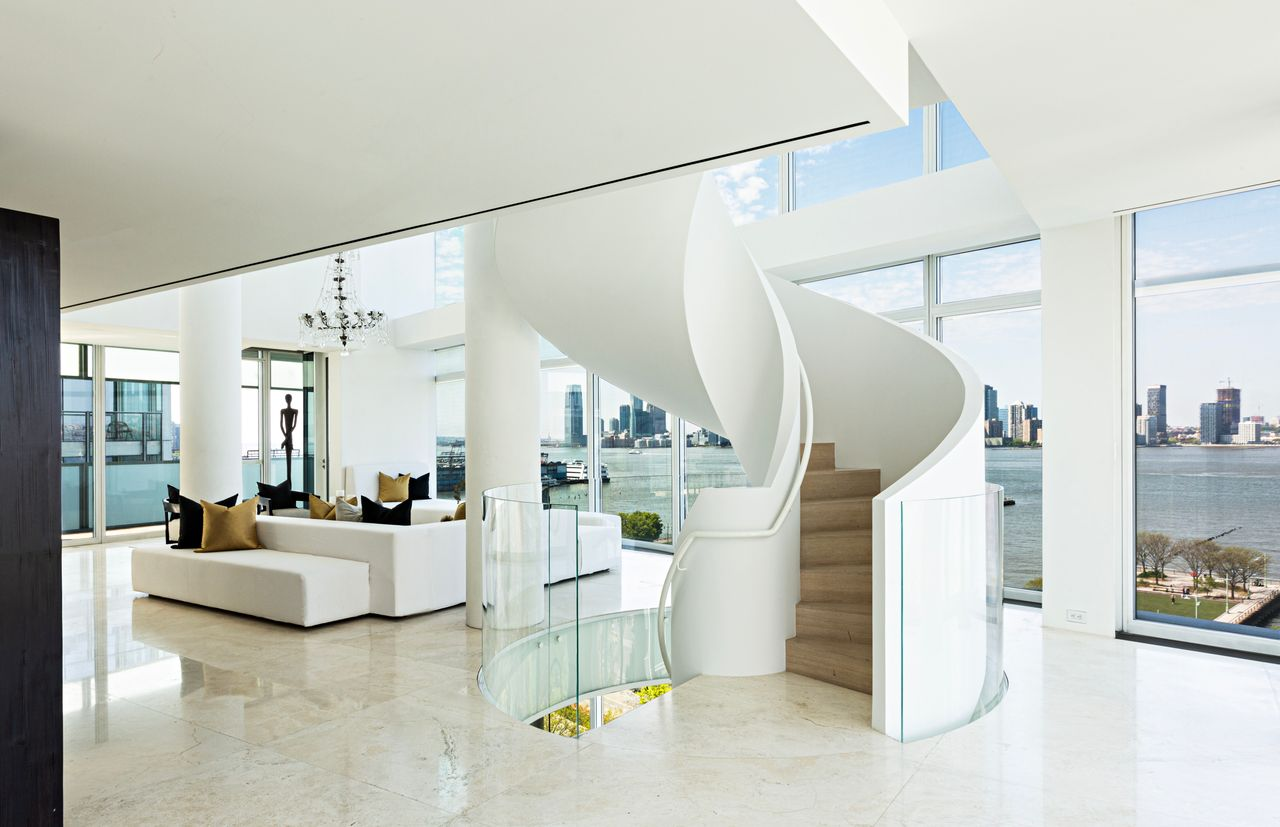 The curved white staircase sits behind the living area, with glass guardrails wrapping the opening, while the space extends forward into low, white seating areas set across pale stone floors.