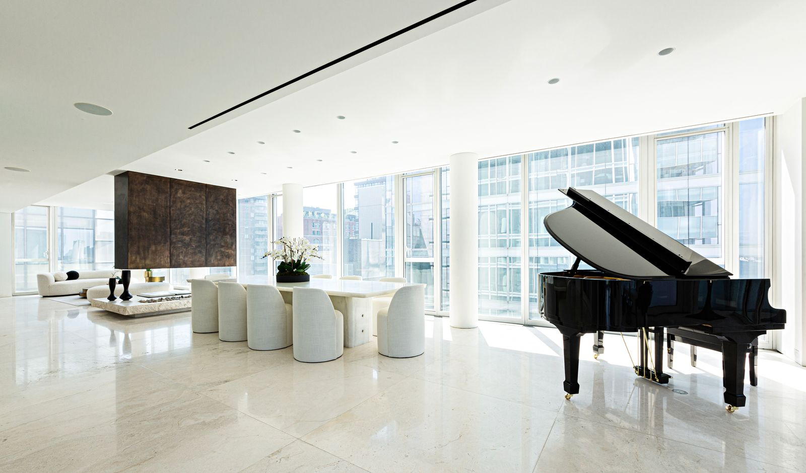 A long white dining table sits near the windows with a row of rounded upholstered chairs gathered around it, while a black grand piano sits to the right.