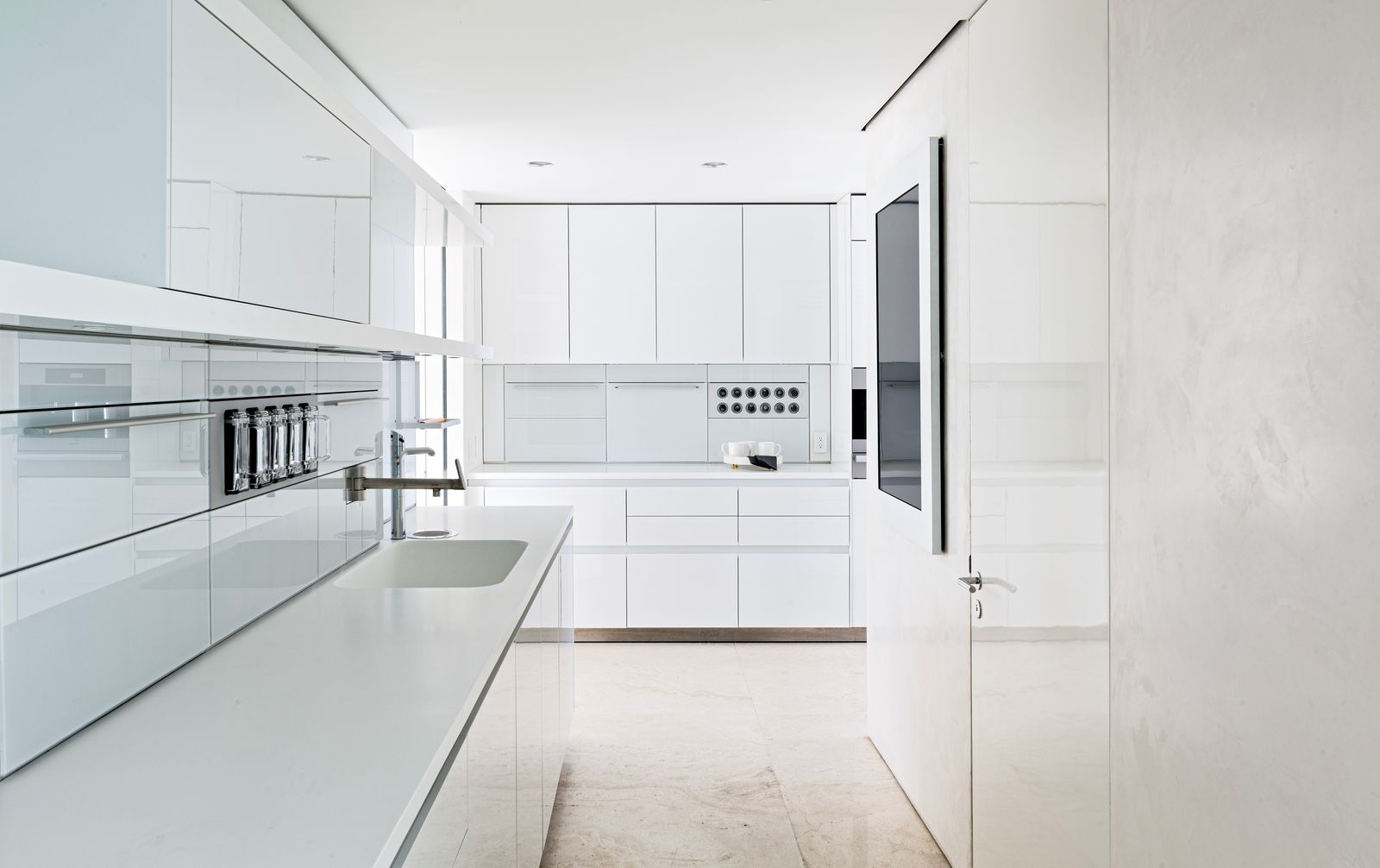 The narrower passage opens into a secondary prep kitchen with cabinetry, walls, and counters all finished in white and built-in appliances tucked behind flat panels for a streamlined look.